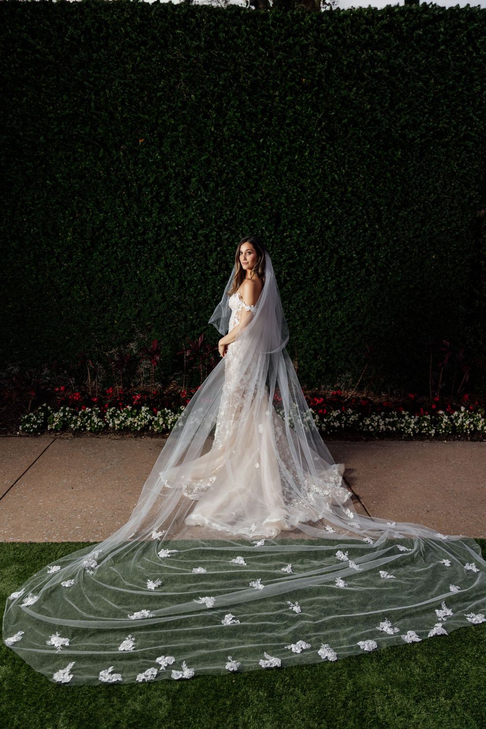 A bride stands in a garden setting wearing an elegant white gown with an extraordinarily long floral-adorned cathedral veil.