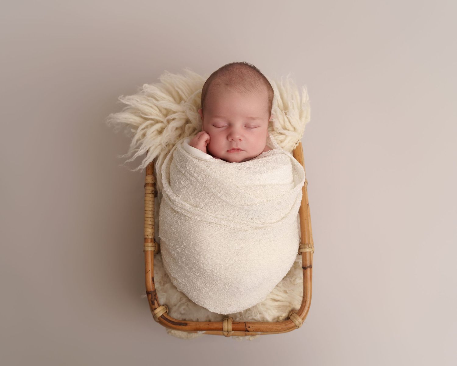Newborn photography of sleeping baby wrapped in white blanket on wooden basket with ivory accents.