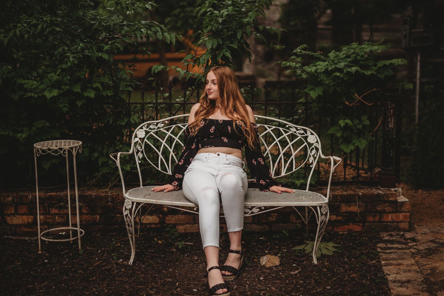 A person in white pants sits on an ornate white metal garden bench against a rustic brick wall background.