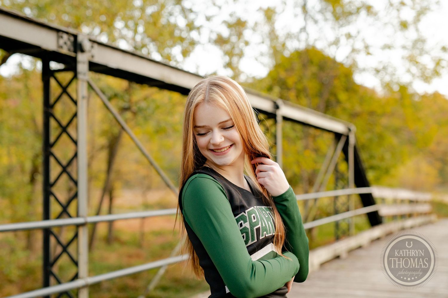 Cheerleader in green varsity uniform smiles warmly while posing on wooden bridge.