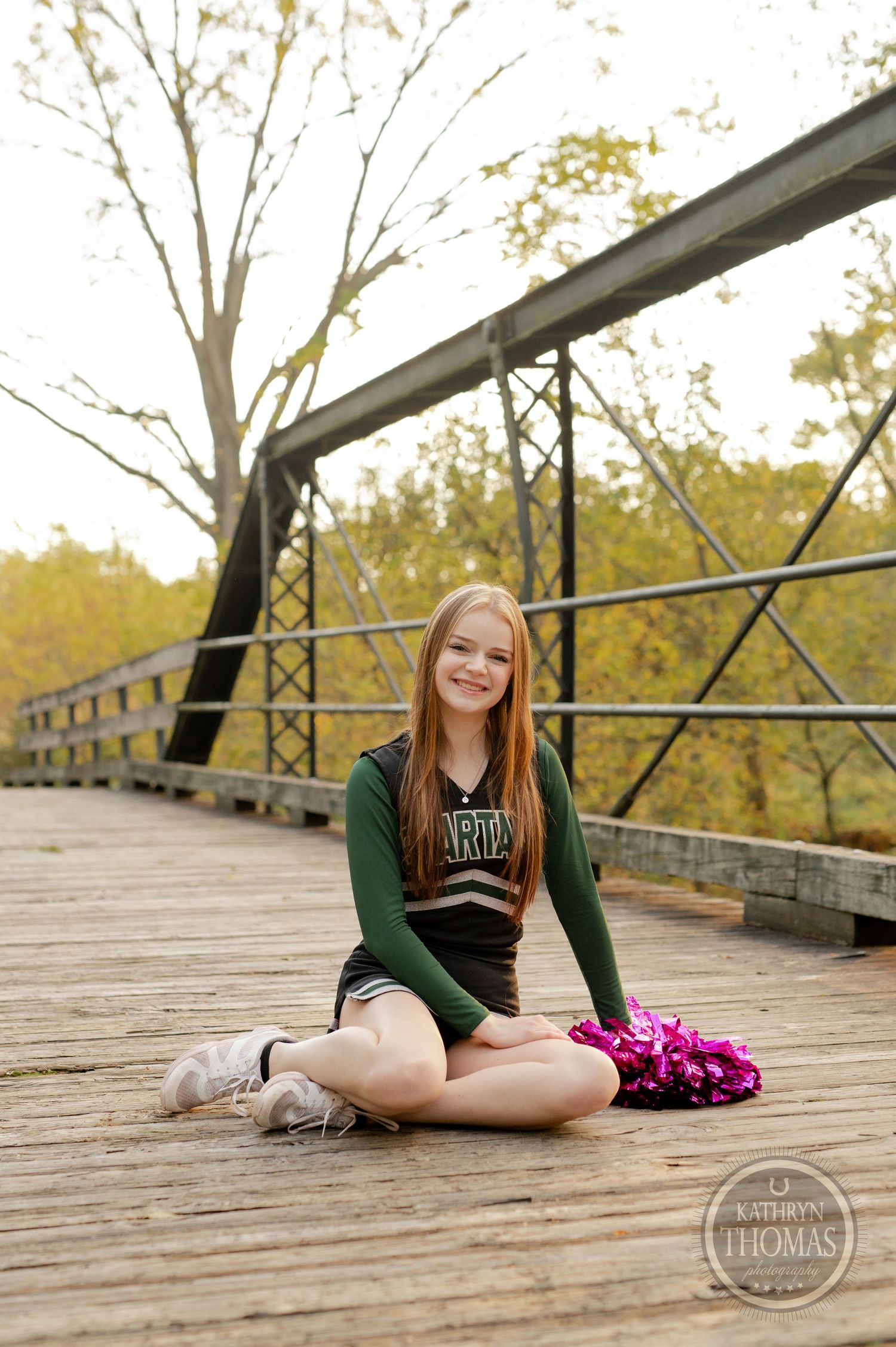 Cheerleader poses sitting cross-legged on wooden bridge with bright pink pom poms and autumn foliage behind.
