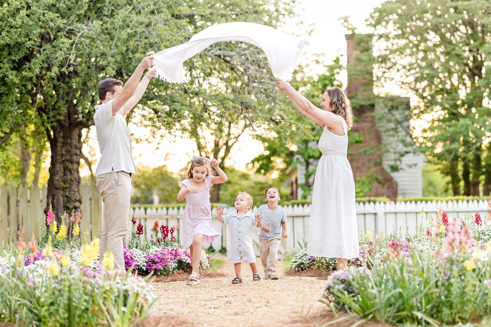 Photography in Colonial Williamsburg of a family playing with a blanket in a cottage garden with flowers and a white picket fence.