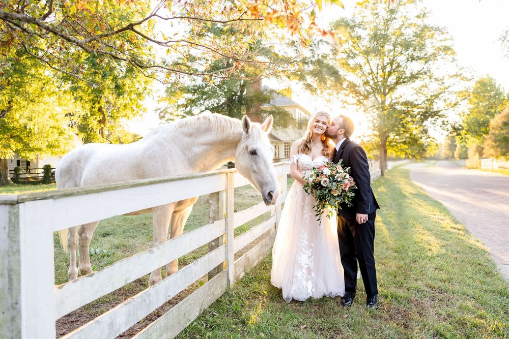 Light and airy wedding portrait in Colonial Williamsburg with a white horse and a white picket fence with sunlight filtering through.