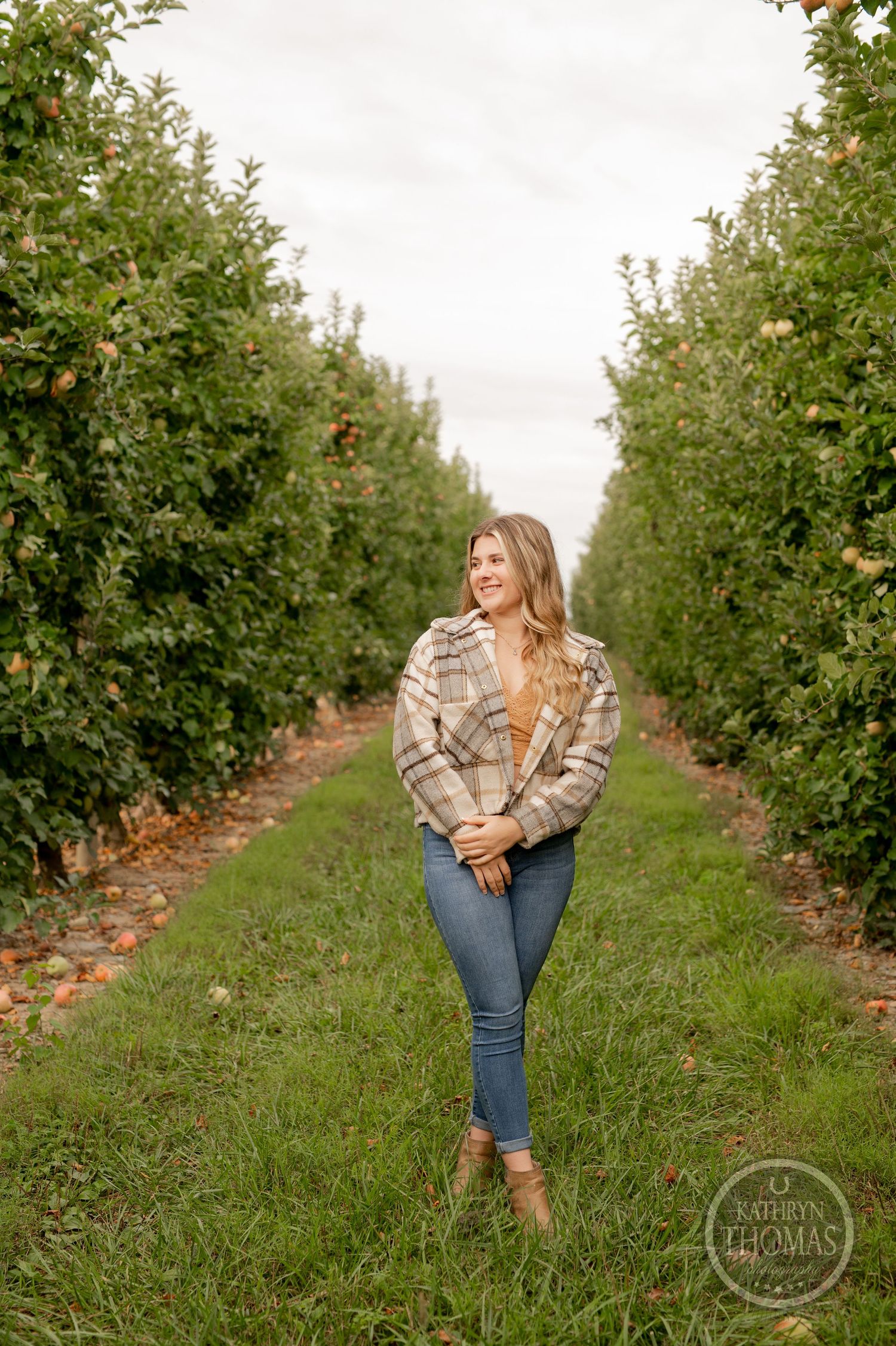 A casual fall portrait showing someone standing between rows of apple trees in a plaid jacket and jeans.