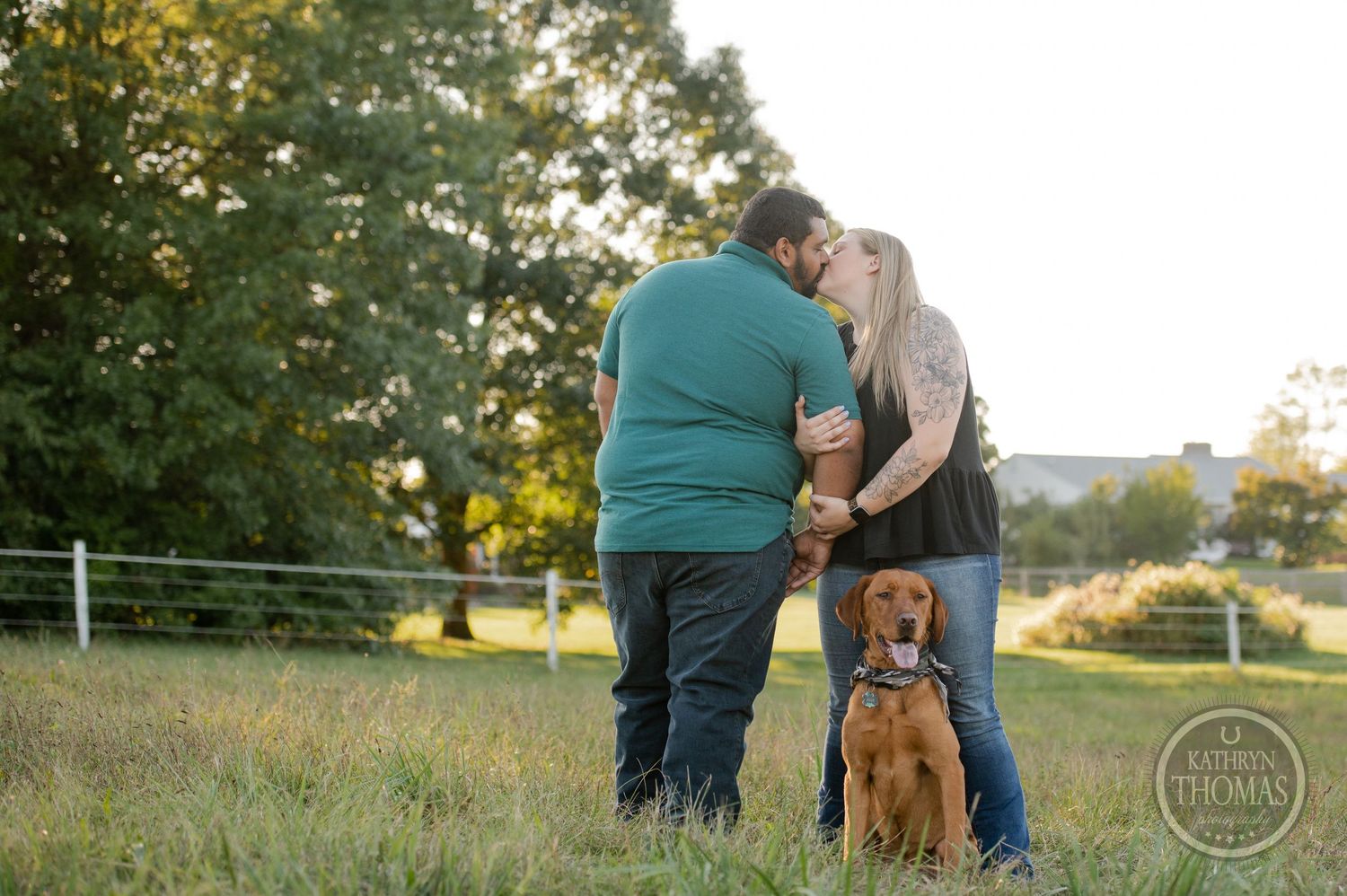 A couple shares an intimate moment in a field while their brown dog sits nearby.