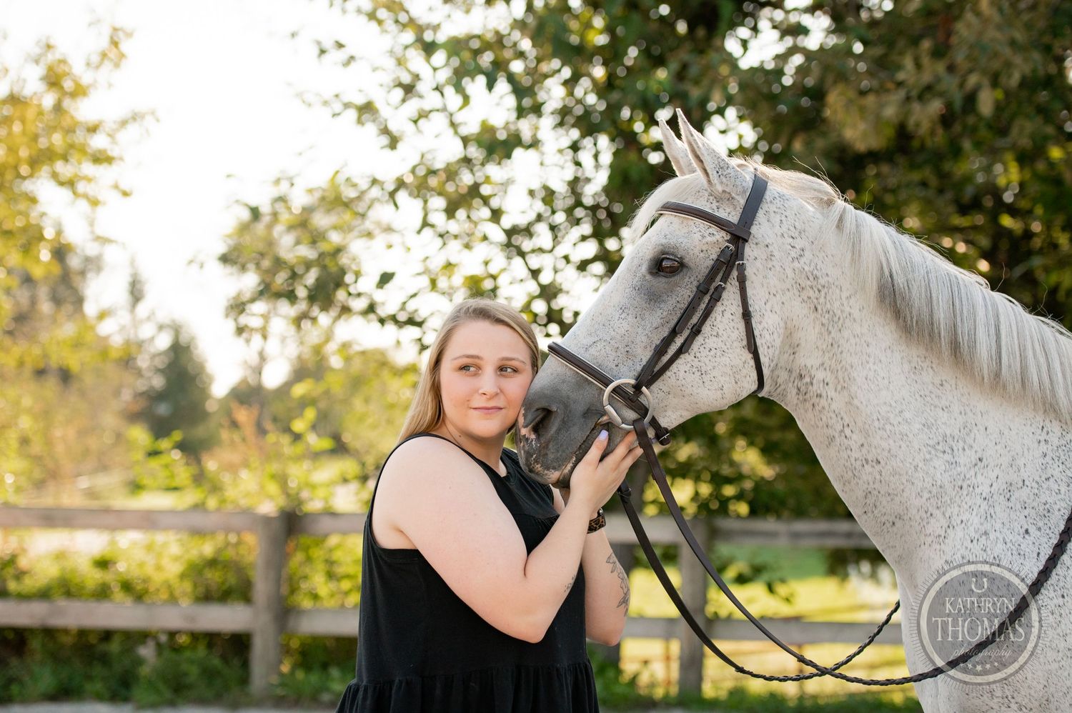A beautiful gray horse and someone in a black tank top pose together in an outdoor paddock at sunset.