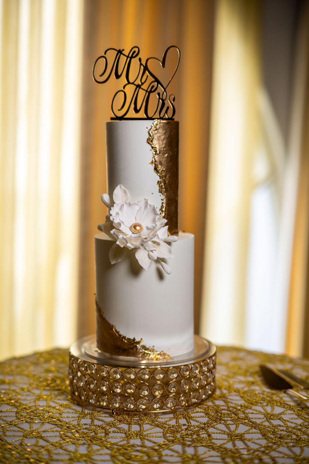 Elegant white and gold wedding cake with 'Mr & Mrs' topper and sugar flower decoration.