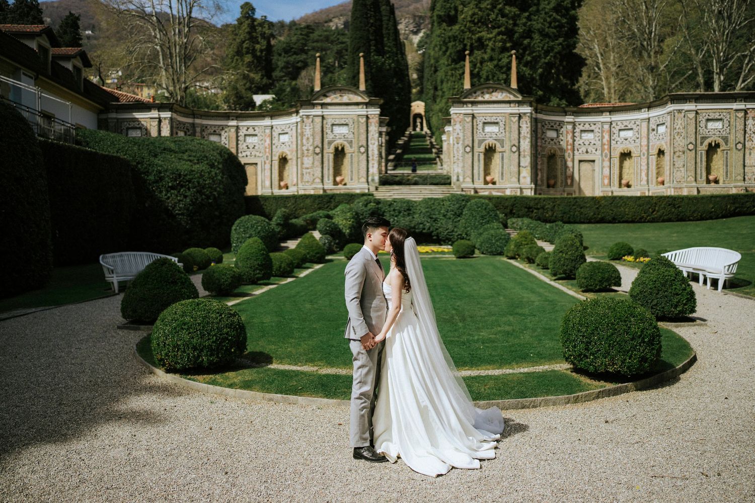 A romantic wedding couple embraces in a formal Italian garden with symmetrical hedges and architecture.