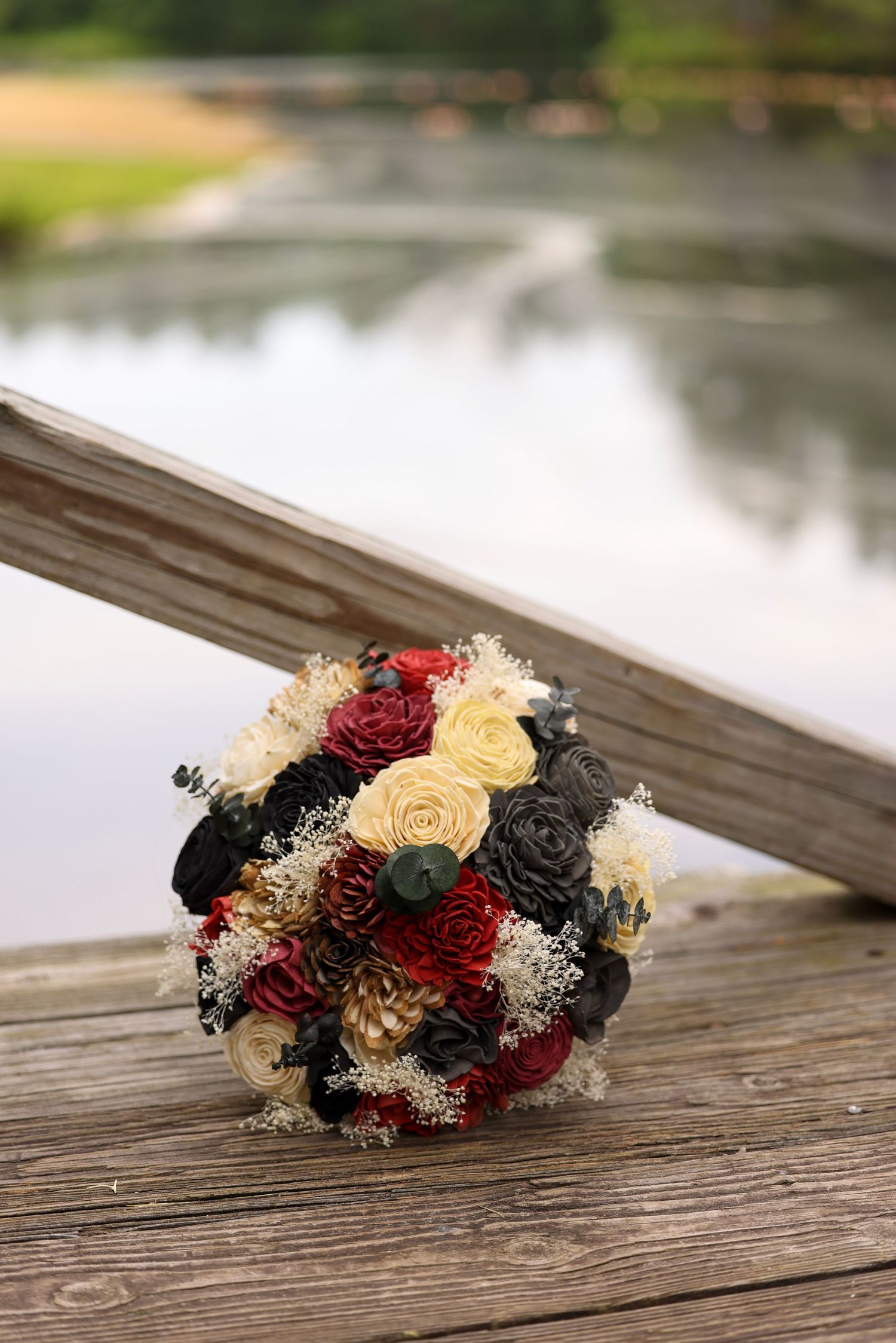 Rustic wedding bouquet with red, cream, and dark roses resting on a wooden dock by water.