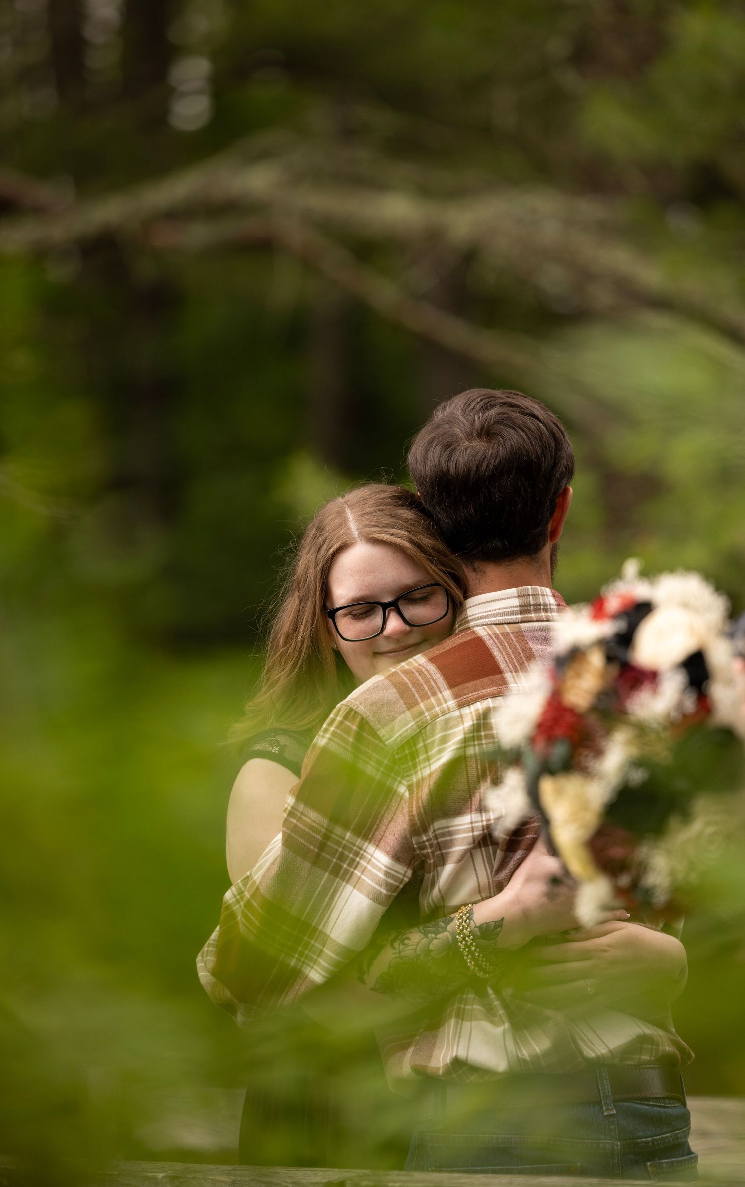 Couple sharing a romantic hug in a lush green forest setting with a colorful bouquet.