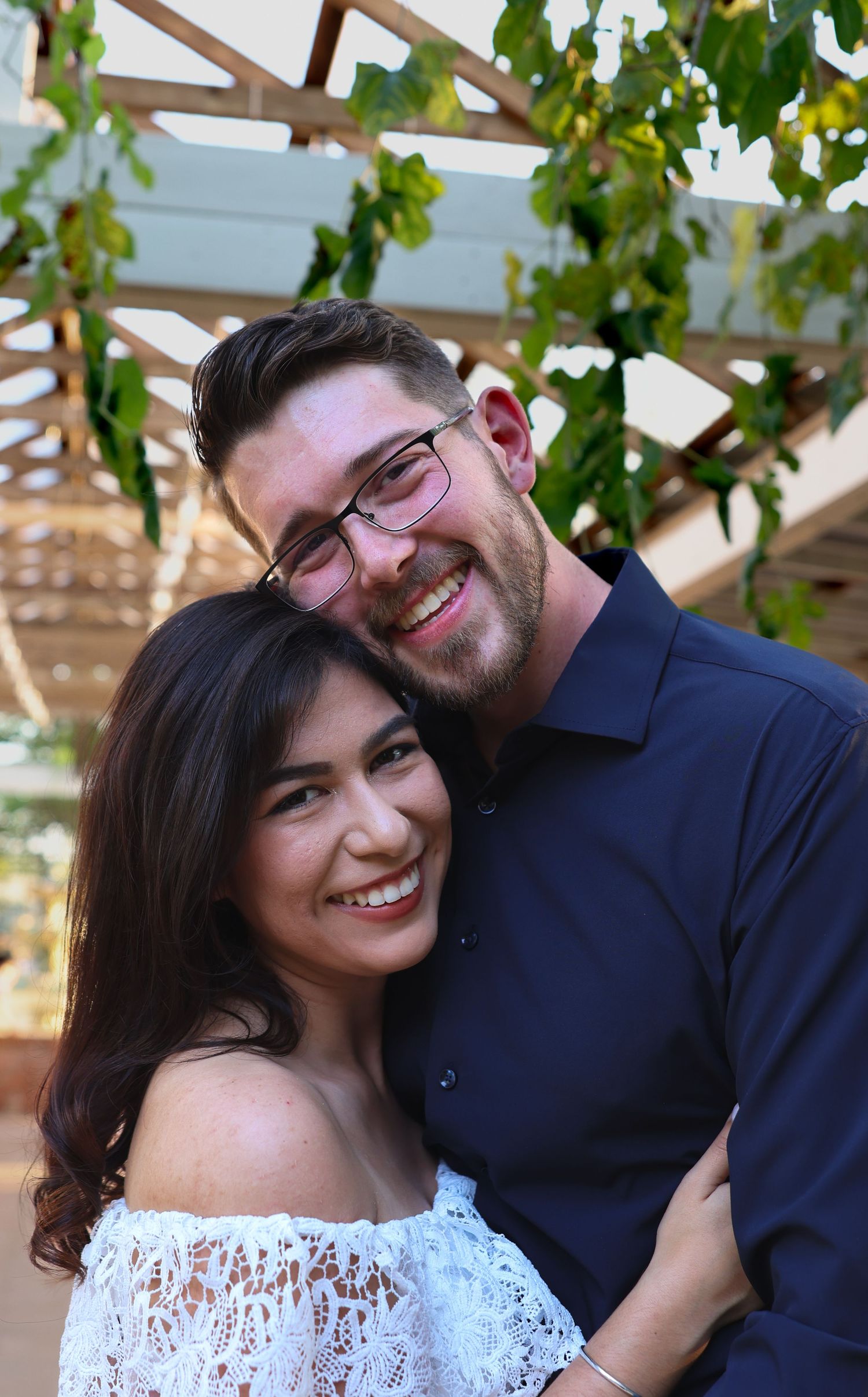 A smiling couple poses together under a pergola with greenery, the woman in a white lace off-shoulder dress.