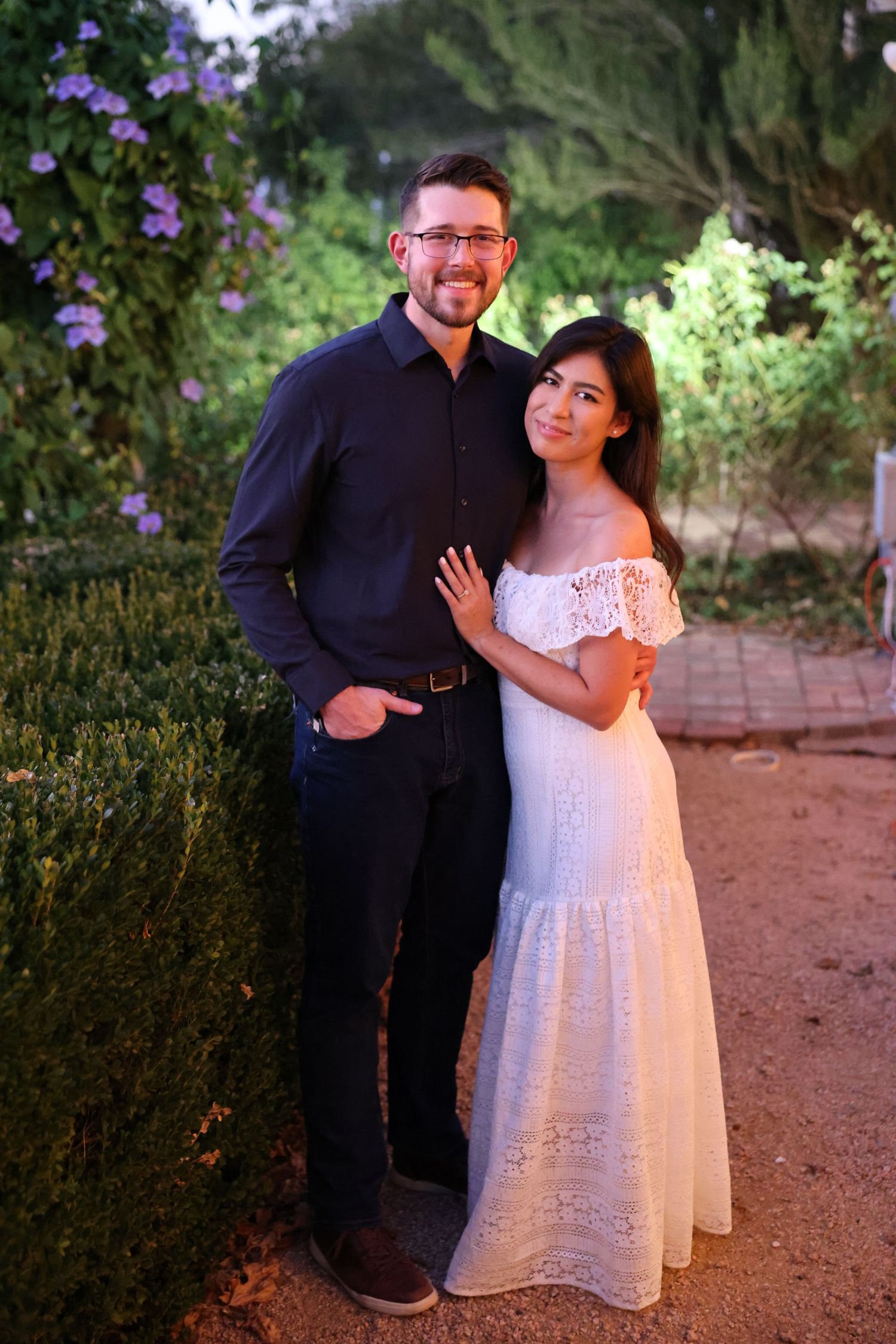 A happy couple poses together along a lush garden path at golden hour, the woman wearing a white maxi dress.