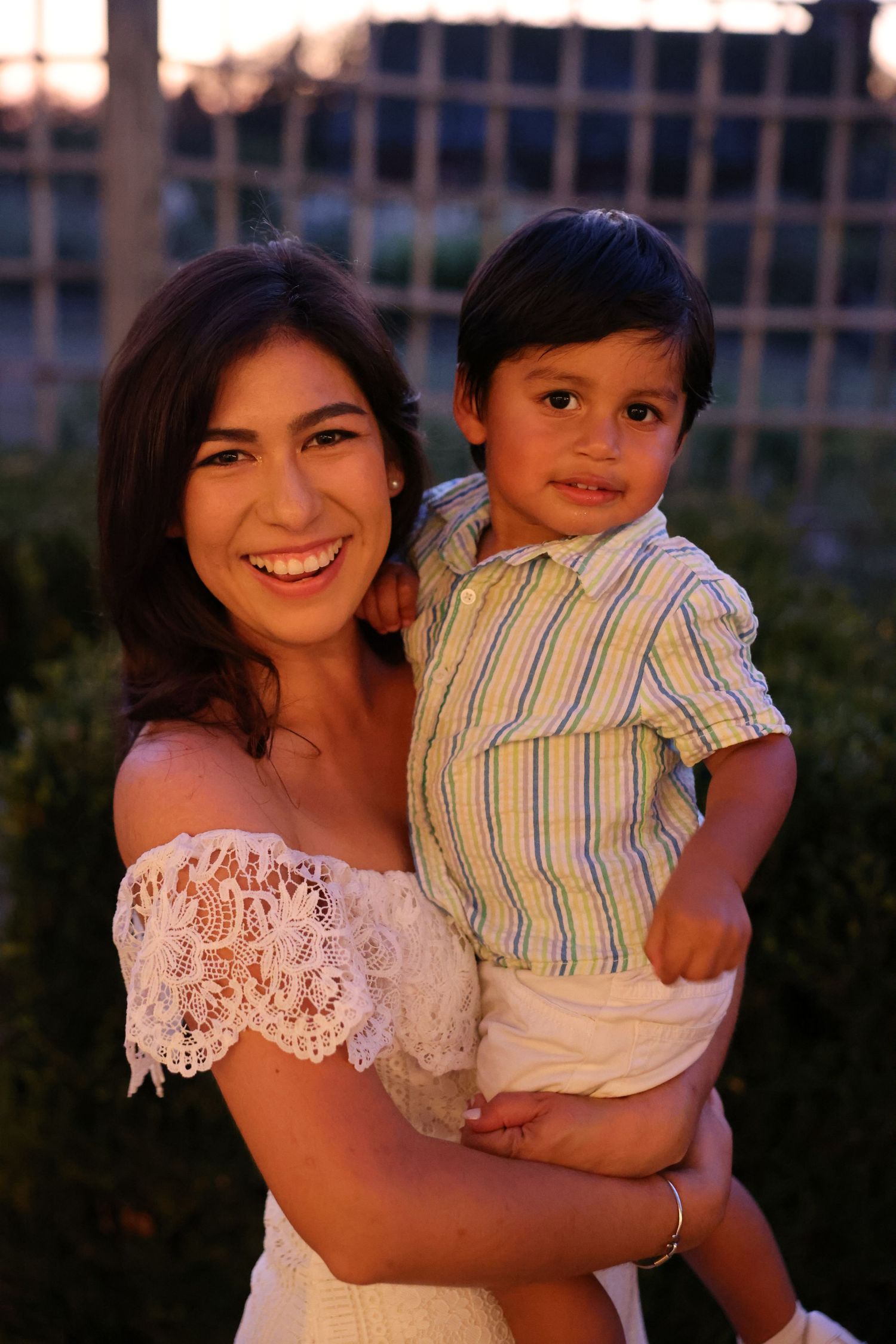 A smiling young woman in a white lace dress holds a happy toddler boy in a striped shirt outdoors.