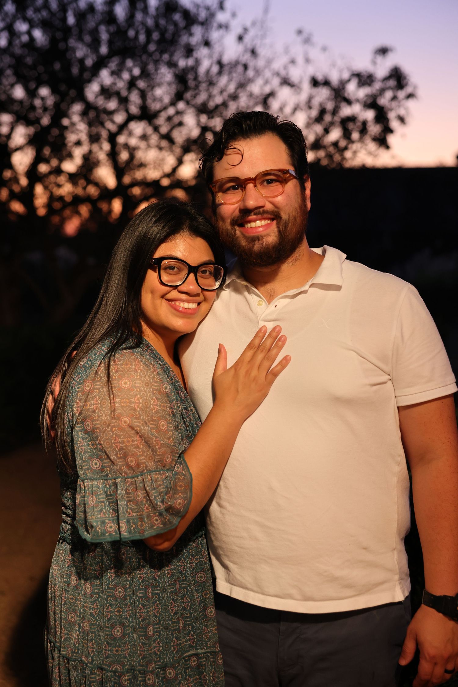 A smiling couple poses together at sunset, the woman showing off her engagement ring to the camera.