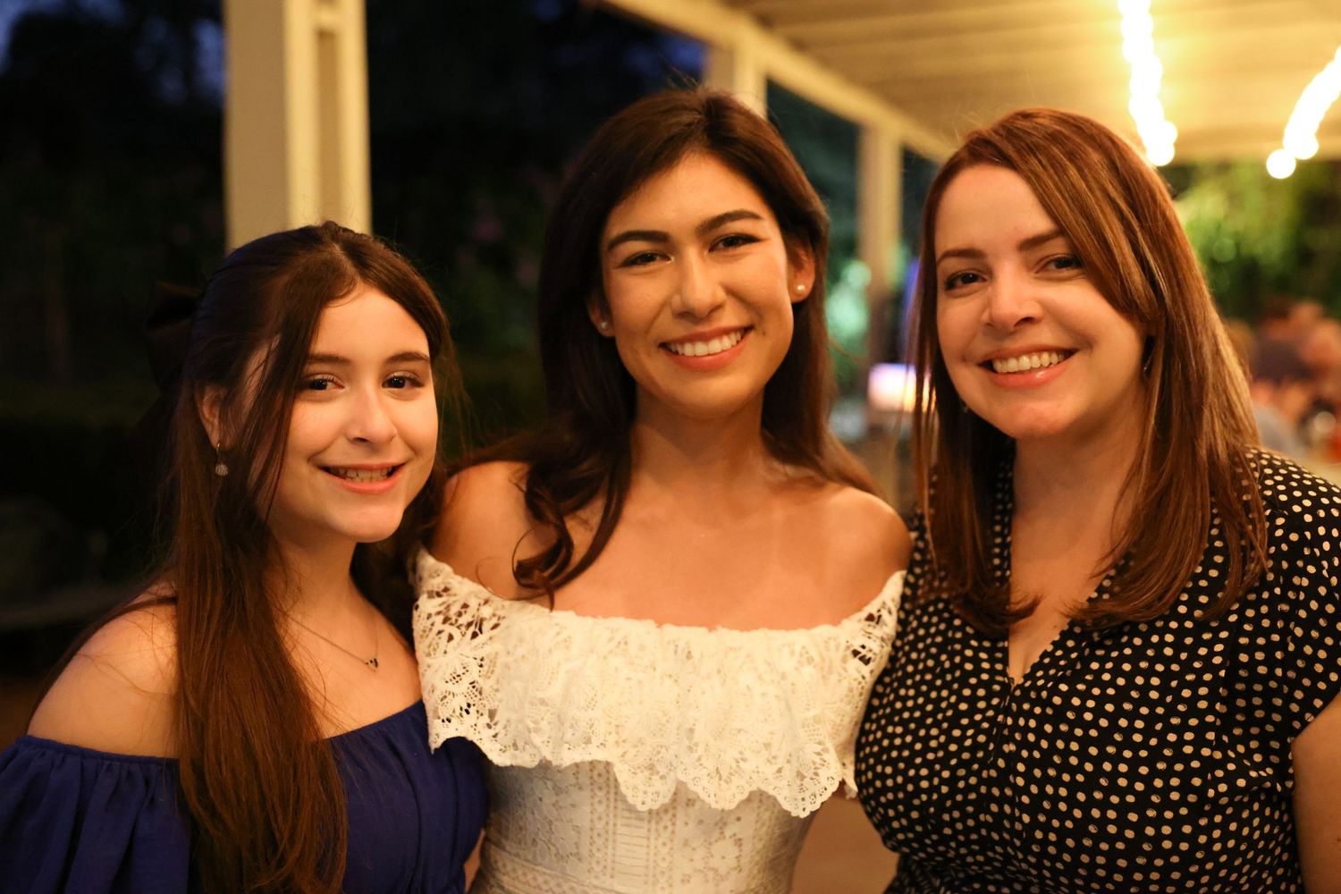 Three women smiling together at outdoor evening celebration.