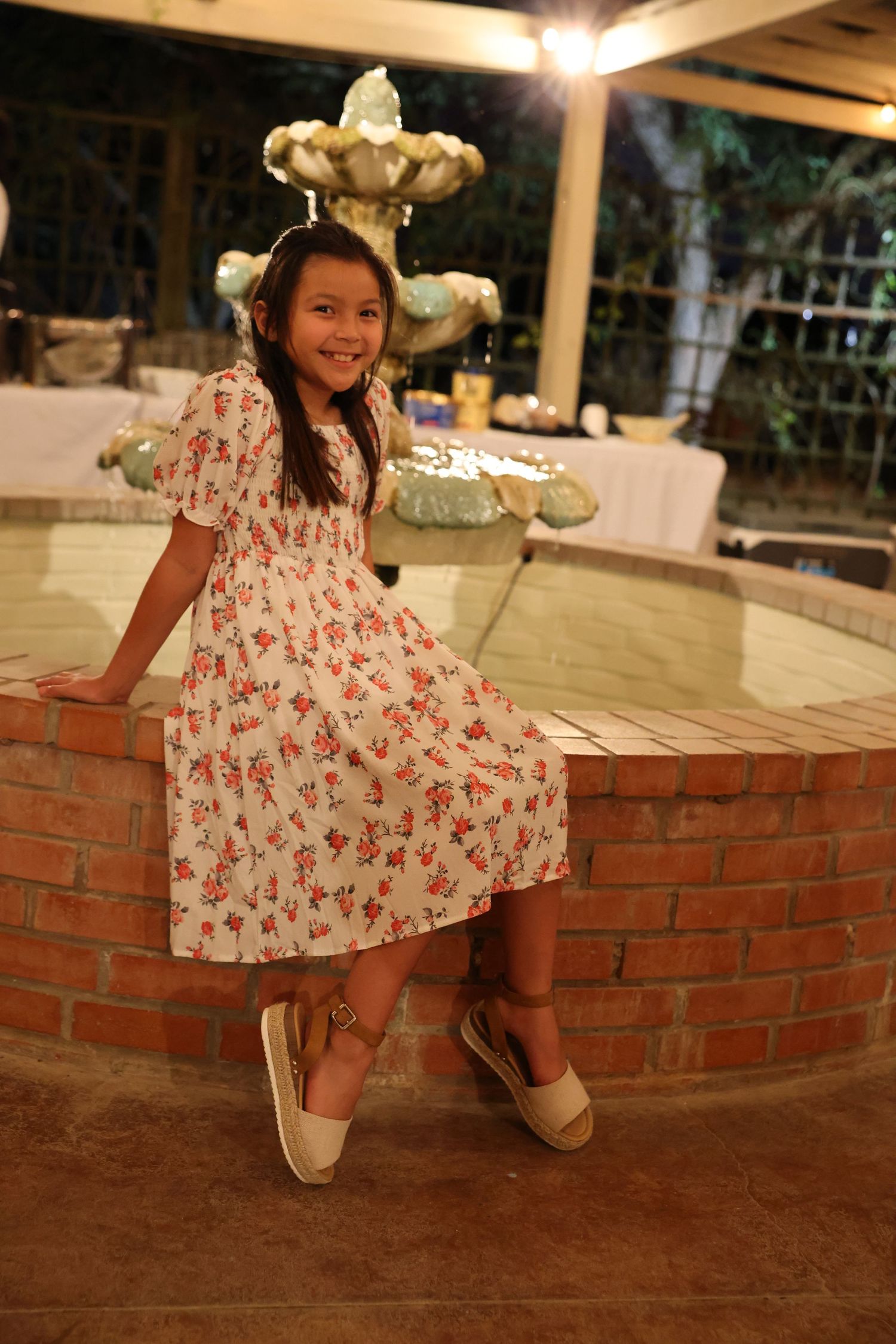 Young girl in floral dress posing by brick fountain at outdoor evening party.