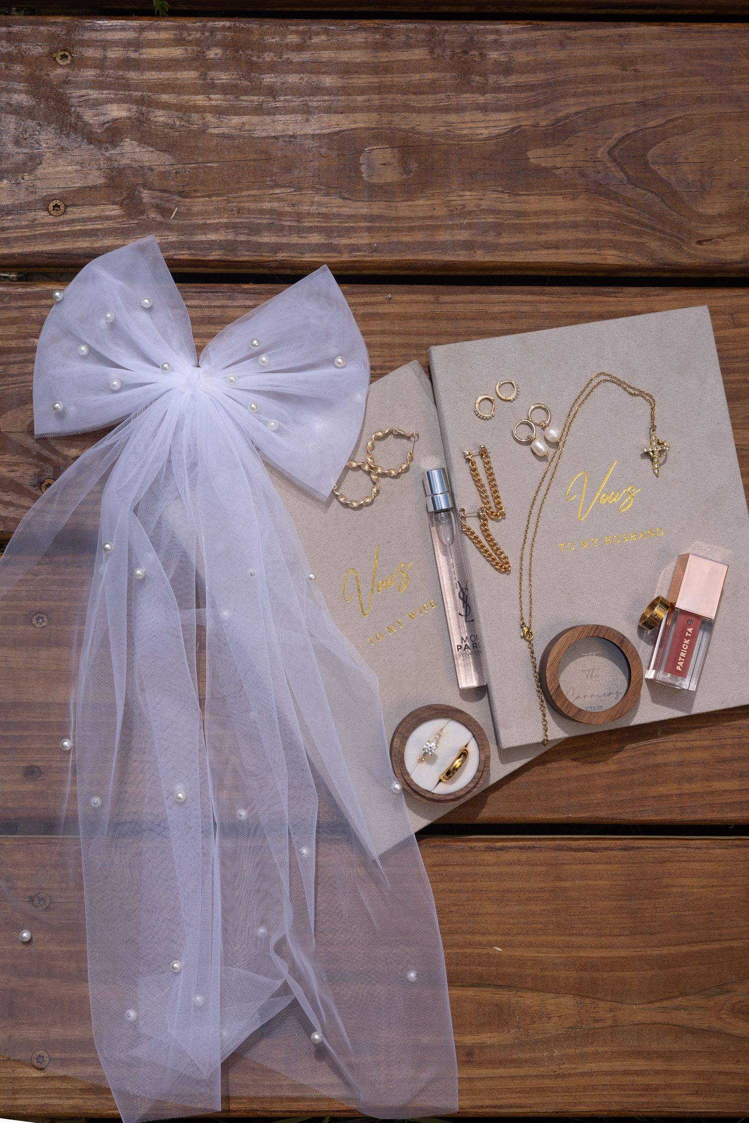 Bridal flat lay featuring pearl veil bow, gold jewelry, perfume, and wedding accessories on wood.