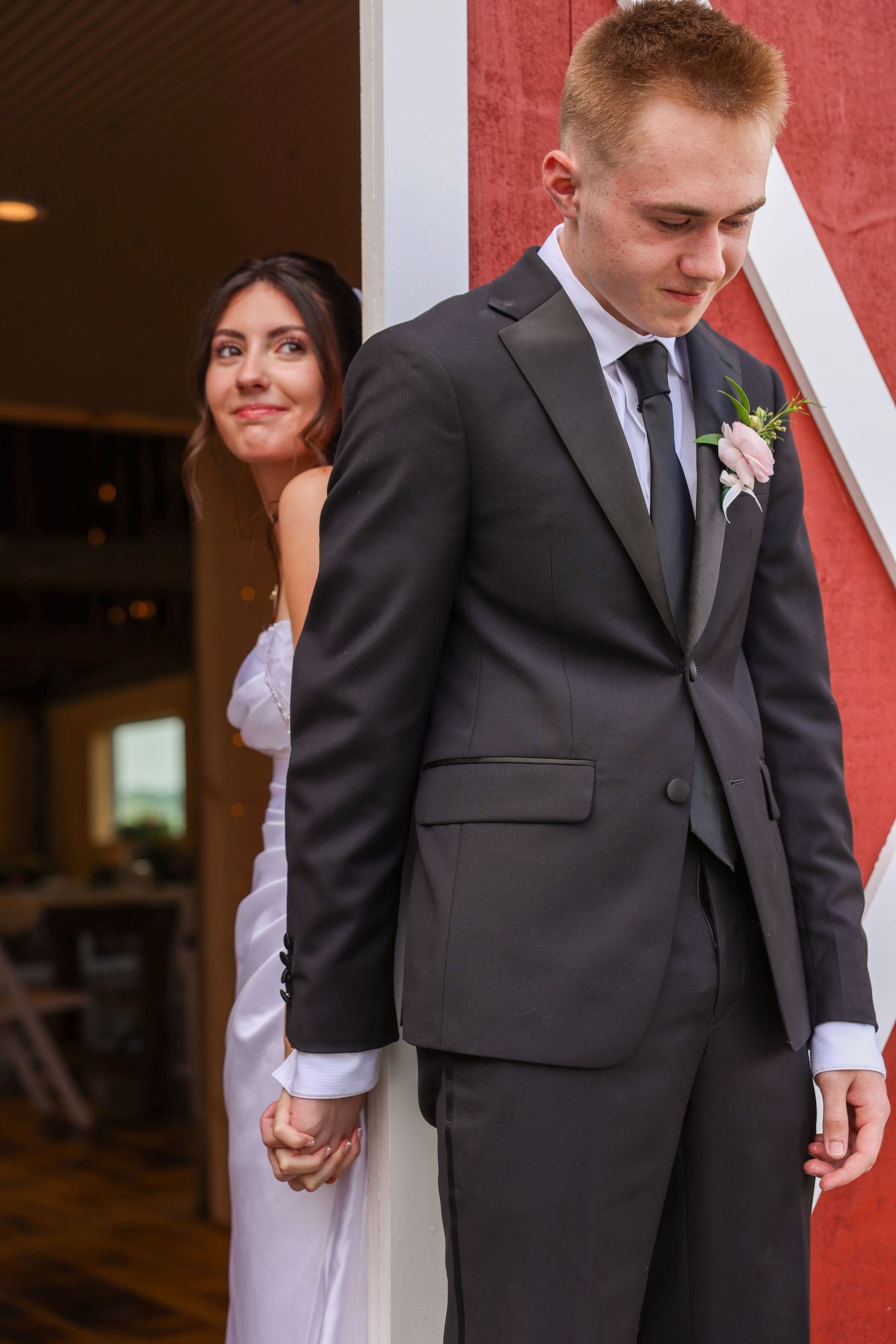 Groom in gray suit holds bride's hand during sweet first look moment at red barn wedding.