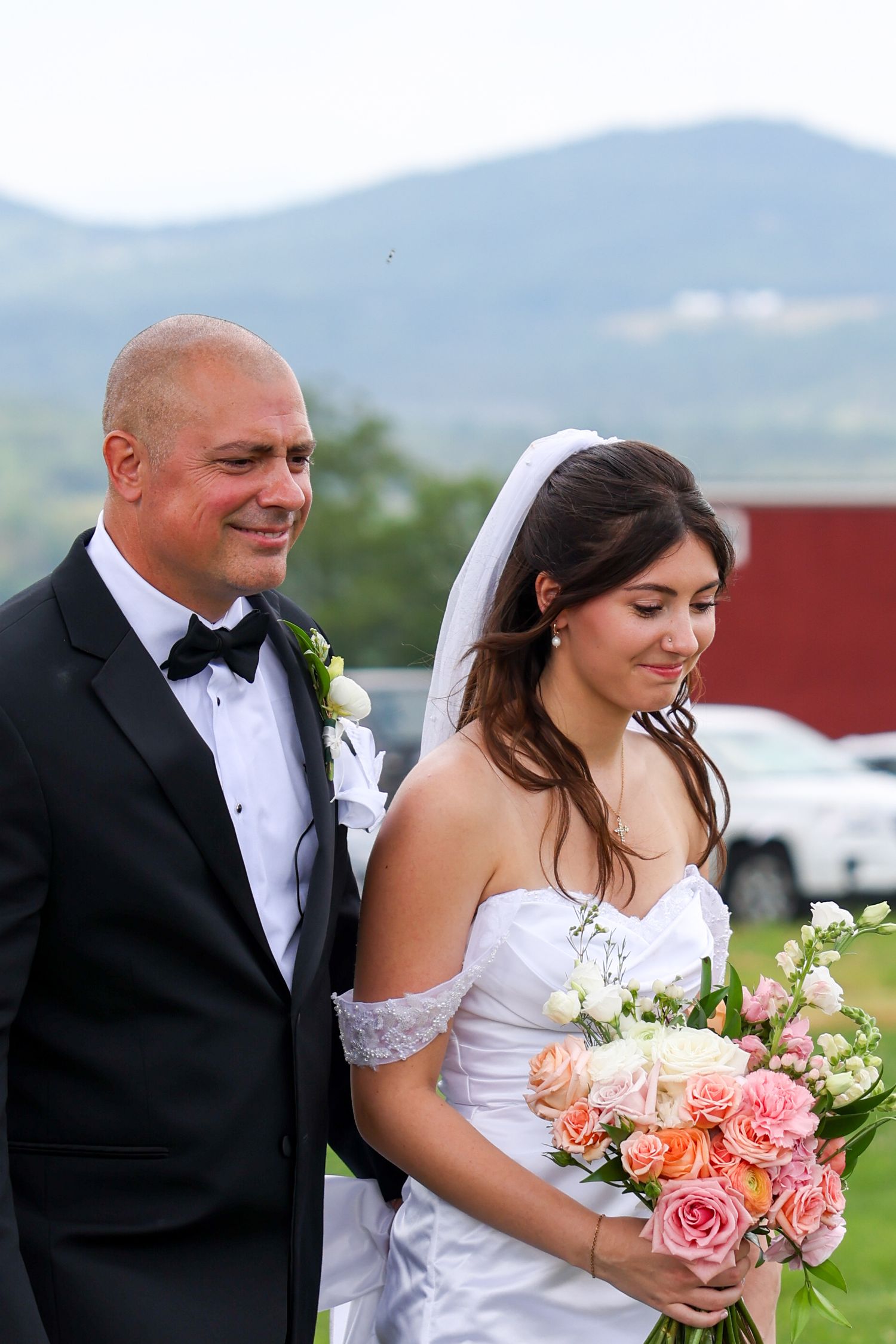 Father walks radiant bride in white gown and veil down the aisle with mountain backdrop.