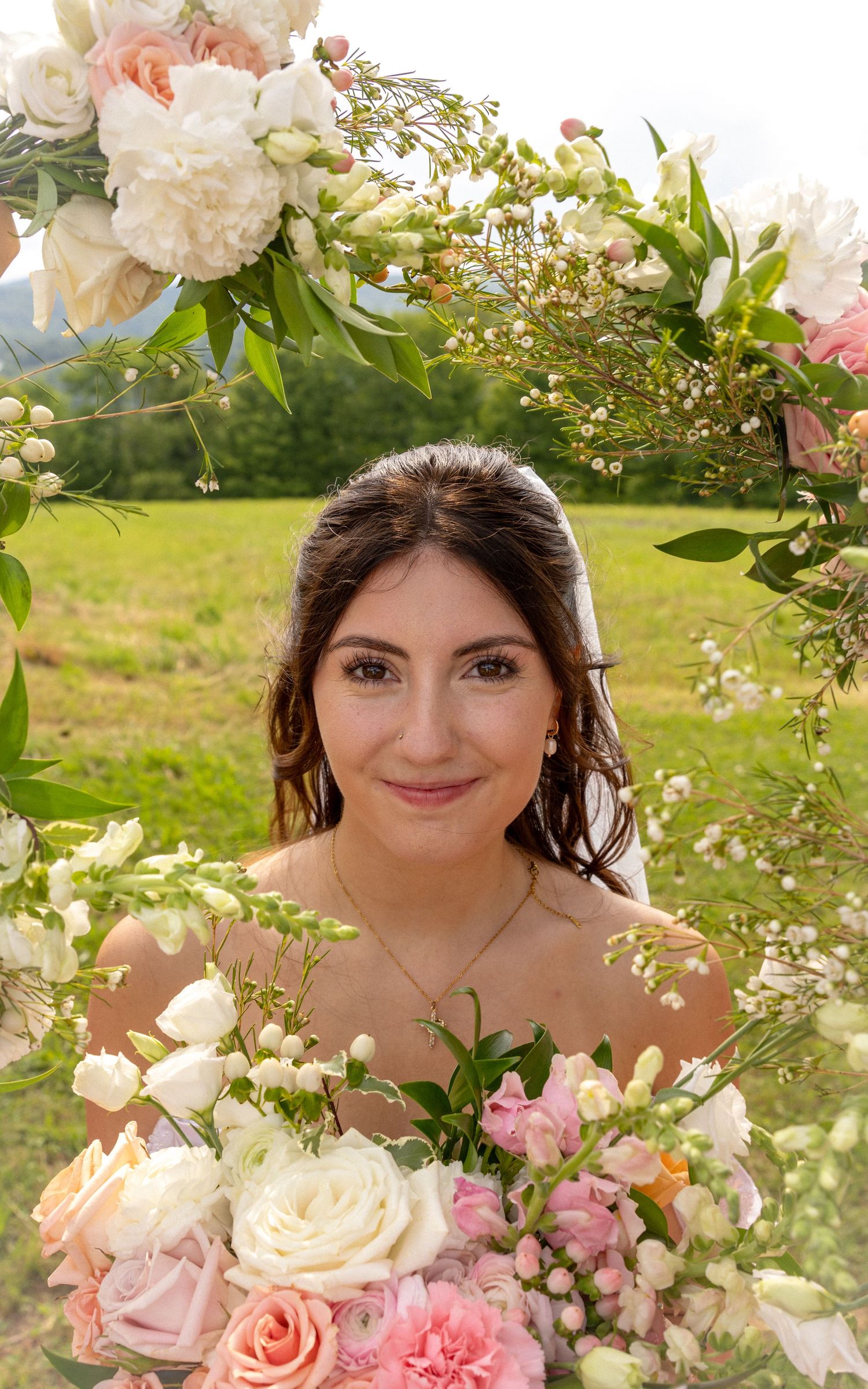 Beautiful bride framed by pink and white floral arch holding matching rose bouquet outdoors.