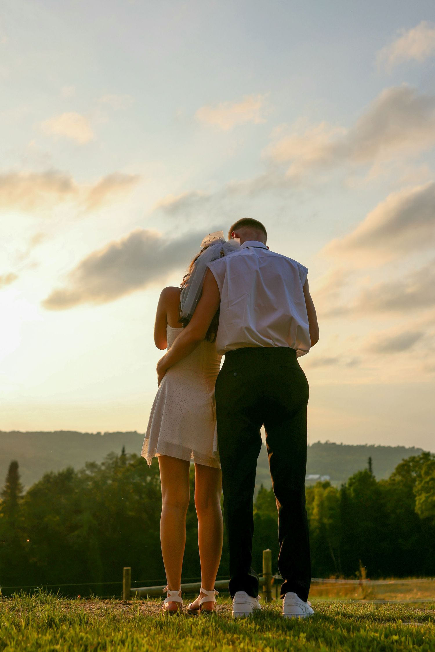 Newlywed couple embraces while watching golden sunset over lush green countryside landscape.
