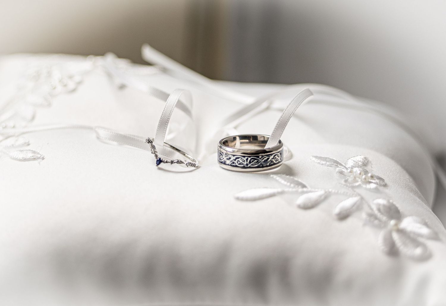 Two elegant wedding bands rest on a white satin ring bearer pillow with delicate embroidered floral details.