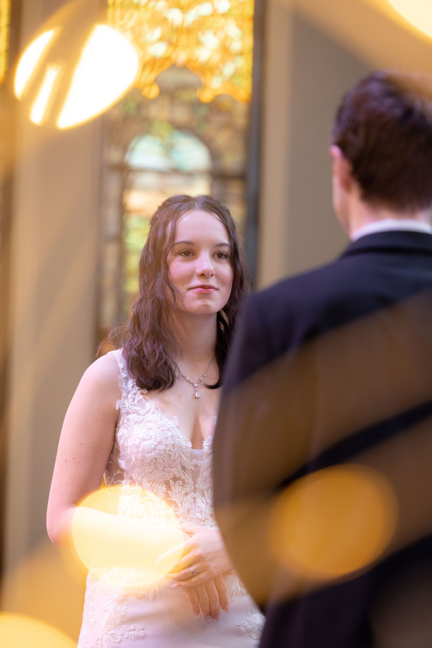 Beautiful bride in lace wedding dress gazing at groom during ceremony with warm golden bokeh lighting.