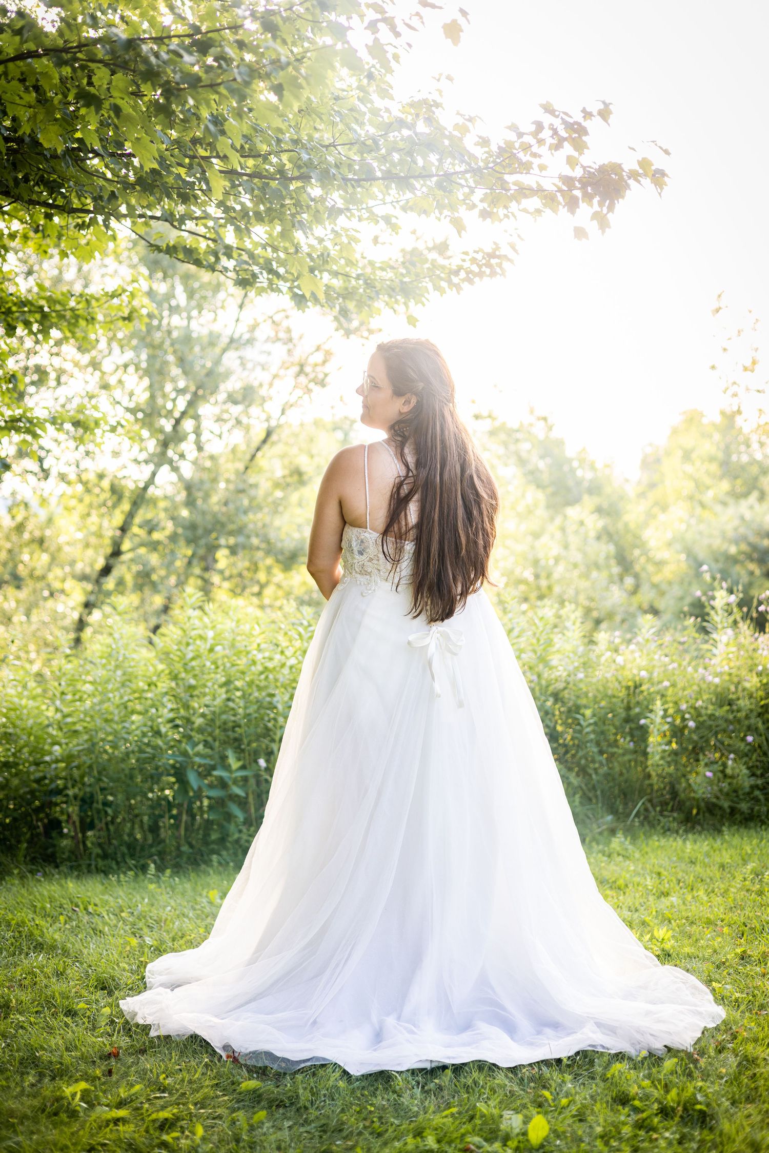Bride in elegant white ball gown with lace bodice stands in sunlit garden, long brown hair flowing down her back.