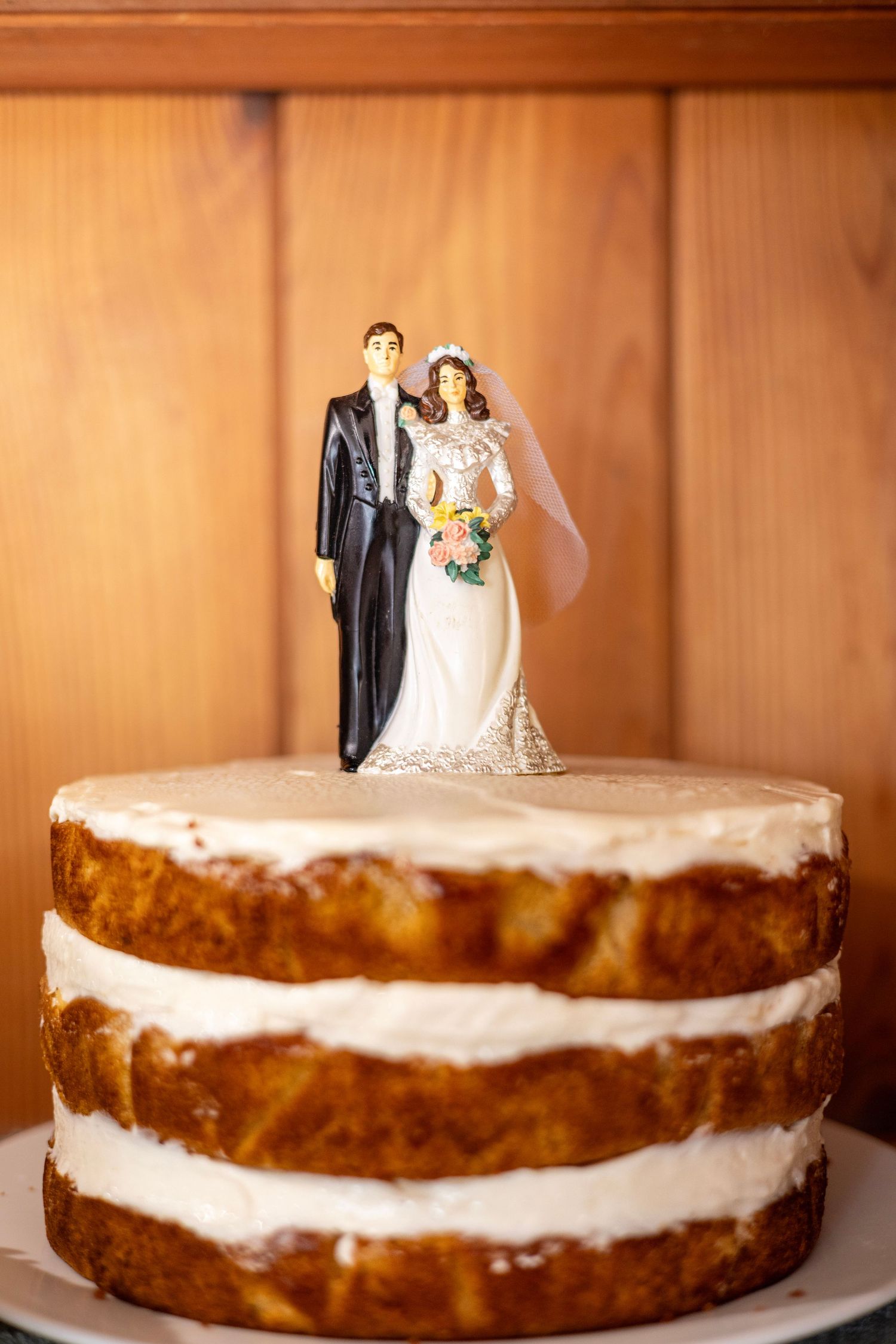 Rustic naked wedding cake with classic bride and groom figurine topper against warm wooden background.