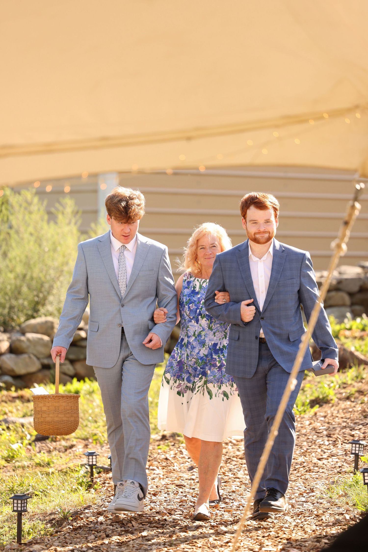 Woman in floral dress walks between two men in suits along a sunlit garden path.