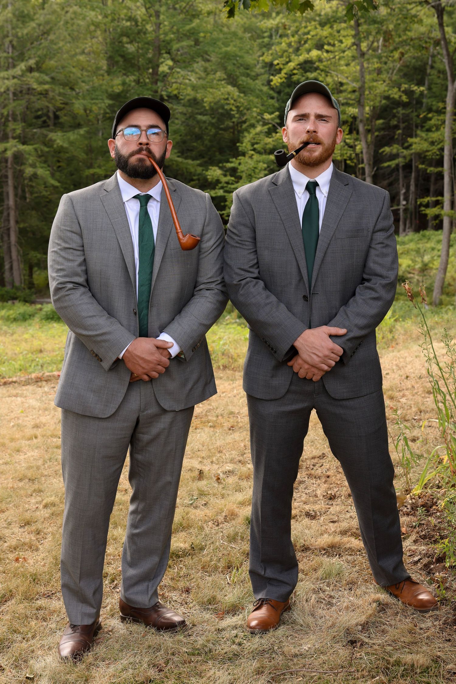 Two men in matching gray suits and caps smoke pipes outdoors in a forest setting.