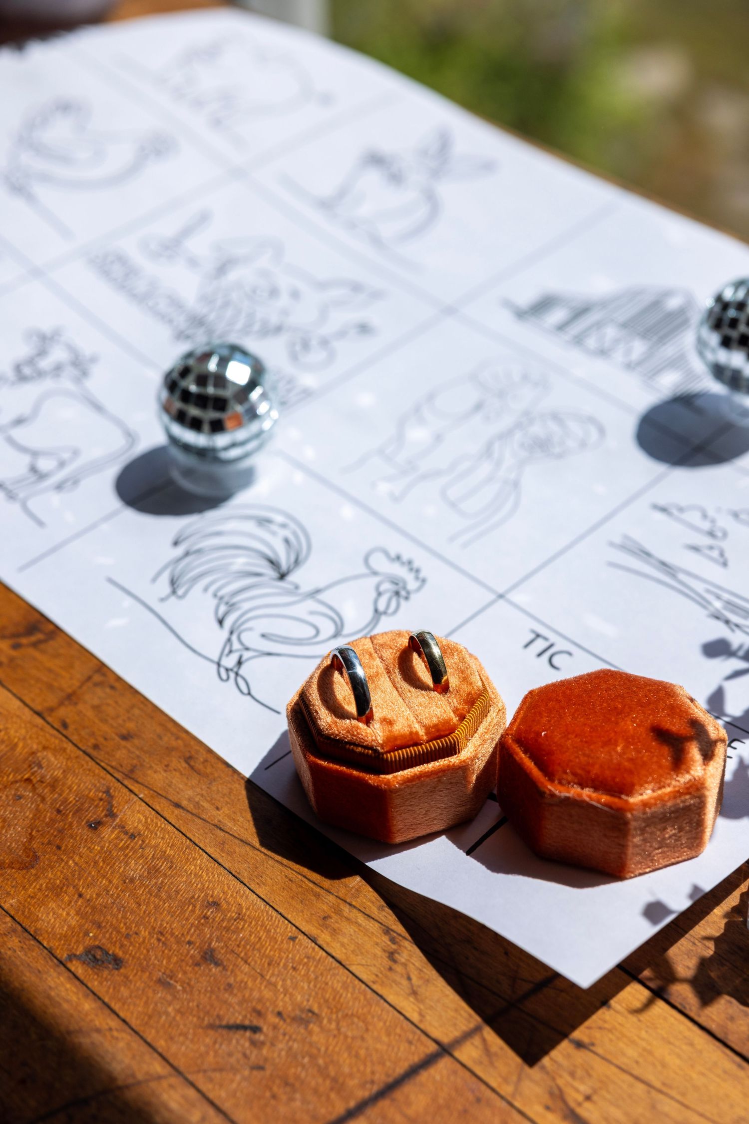 Two wedding bands in orange velvet ring boxes displayed on illustrated paper with mini disco balls on wooden table.