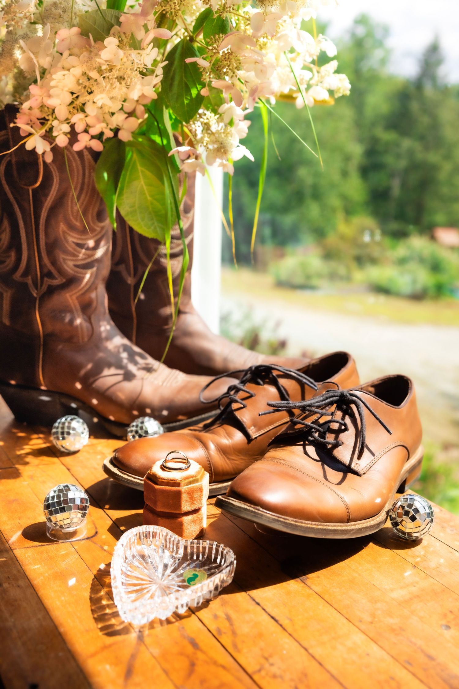Brown cowboy boots and leather dress shoes with wedding rings, disco balls, and white flowers on wooden surface.