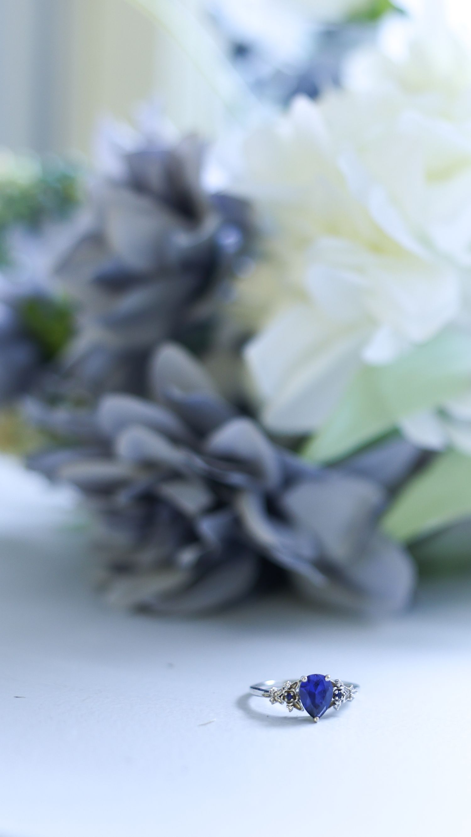 A stunning blue sapphire heart-shaped engagement ring displayed on a white surface with soft floral bokeh background.
