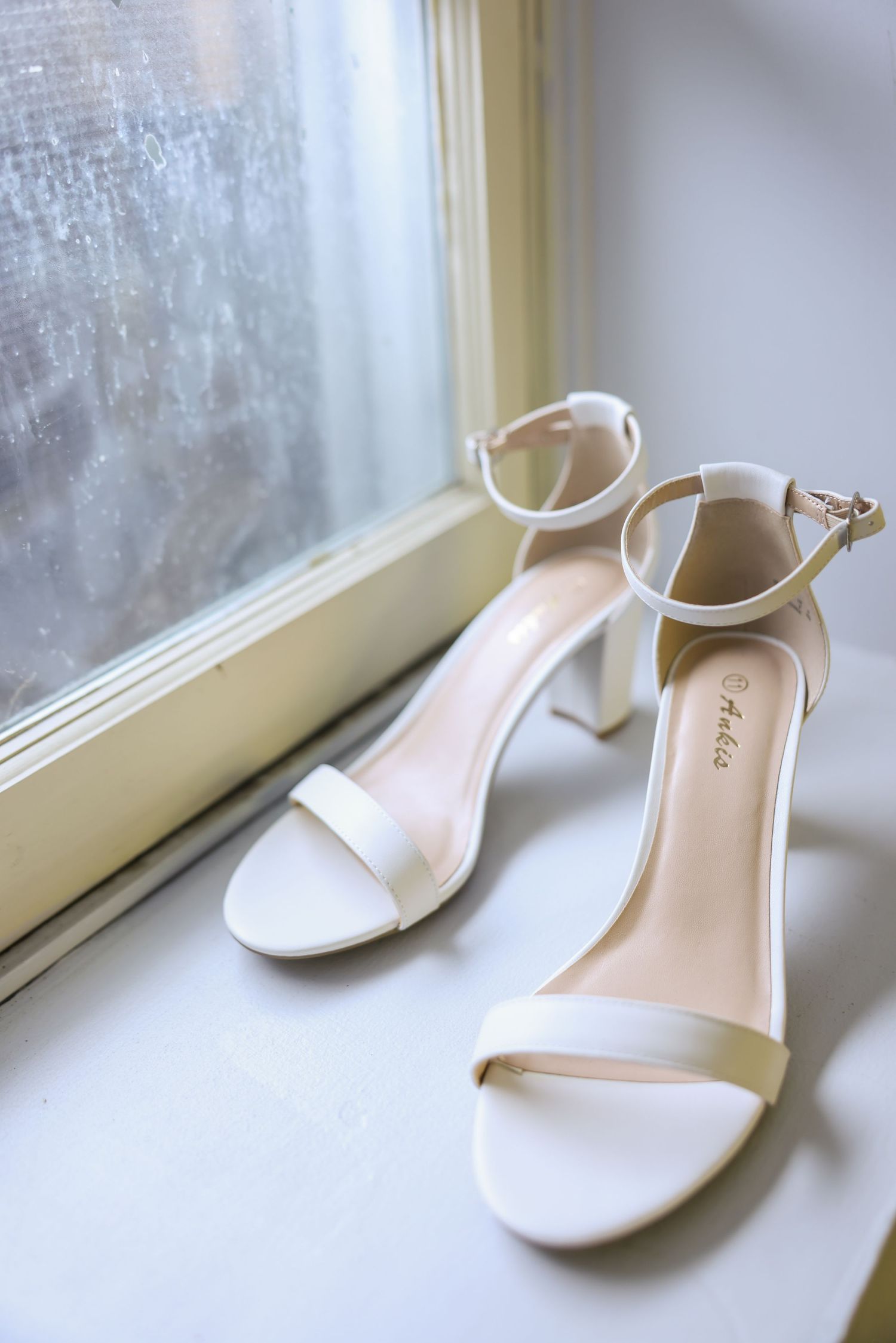 Elegant white block-heel bridal sandals with ankle strap displayed on a window ledge.