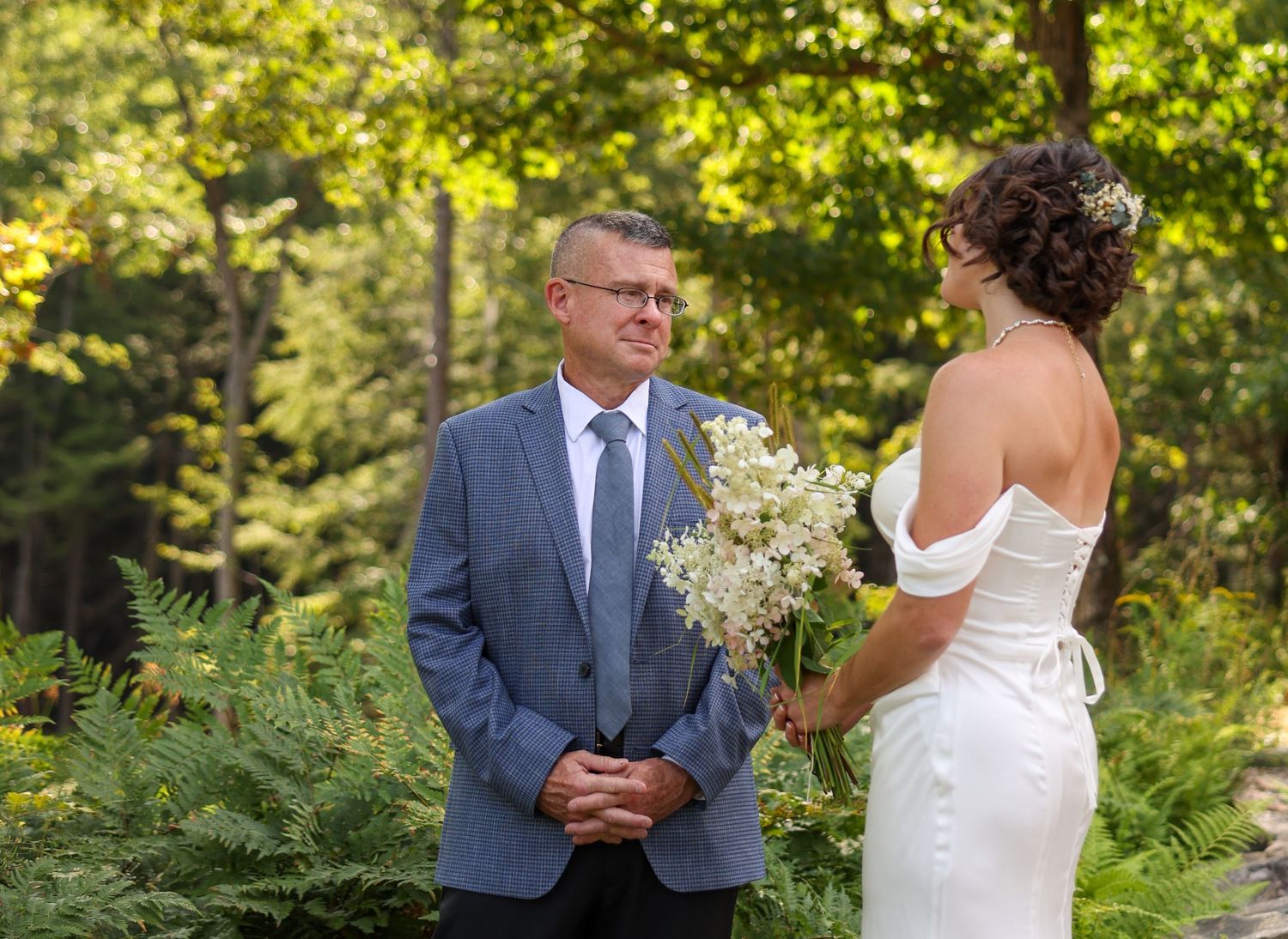 Bride in white gown holds bouquet facing man in blue suit during outdoor wedding ceremony.