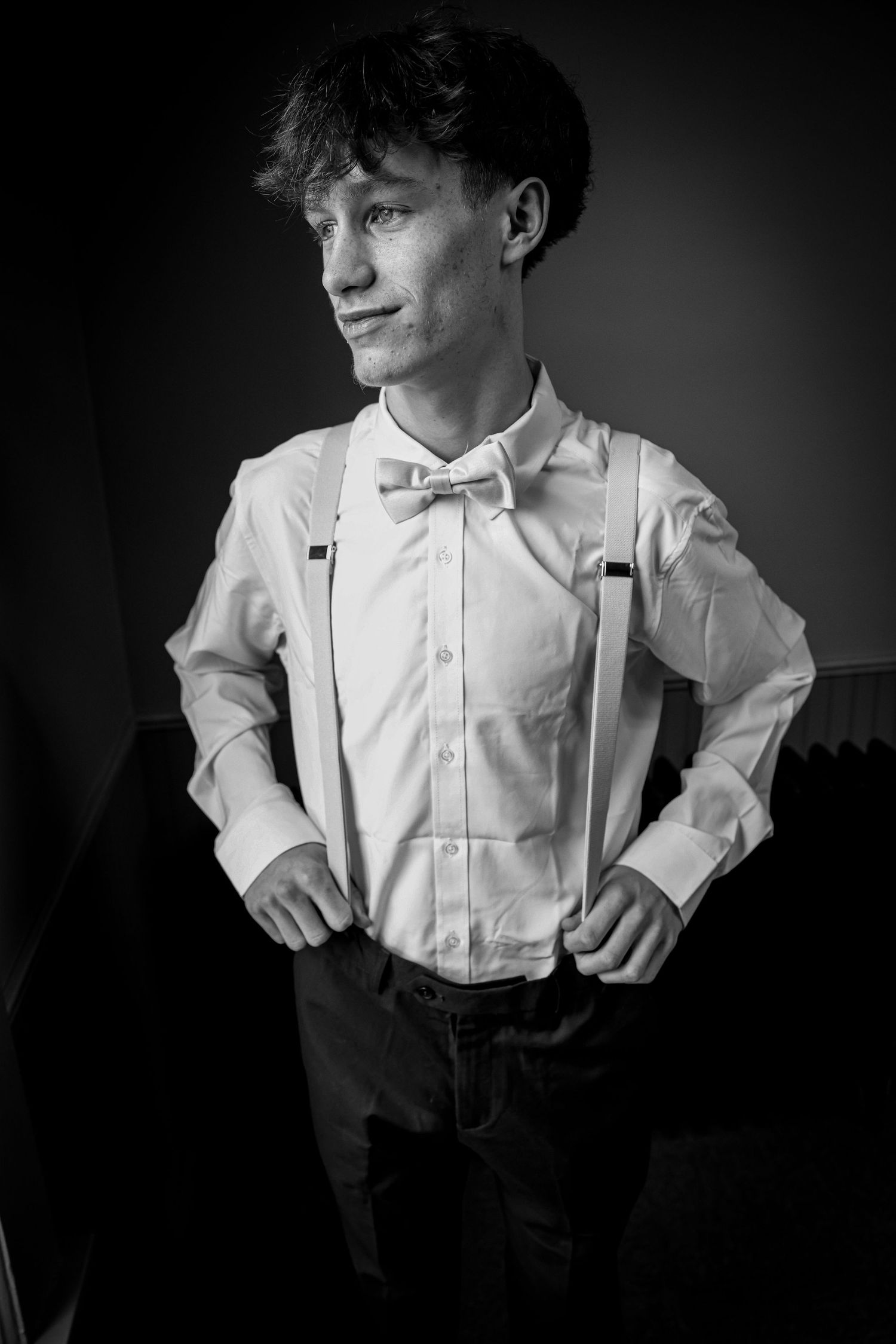 Stylish young groom wearing white dress shirt with bow tie and suspenders in a dramatic black and white portrait.