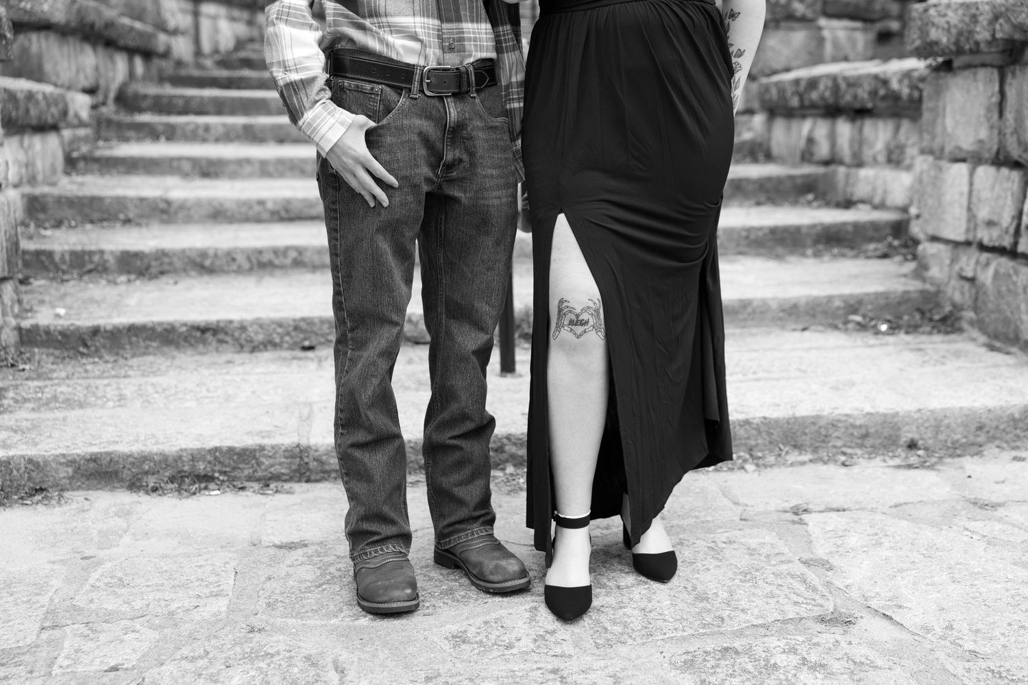 Black and white photo of couple standing on stone steps showing jeans, boots, and black dress.