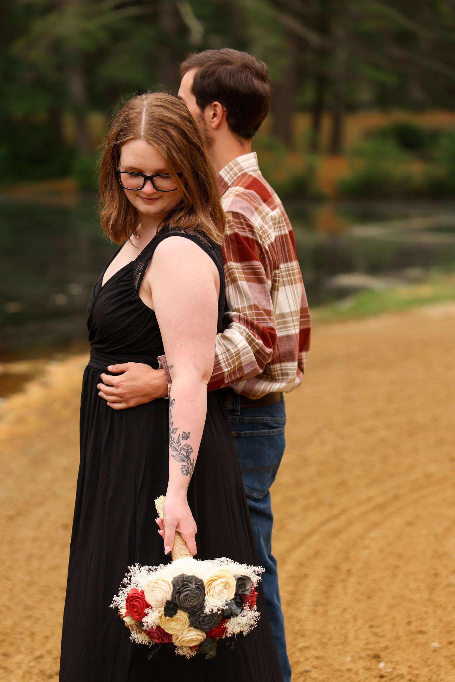 Woman in black dress with tattoos holds bouquet while embracing partner near a sandy riverbank.