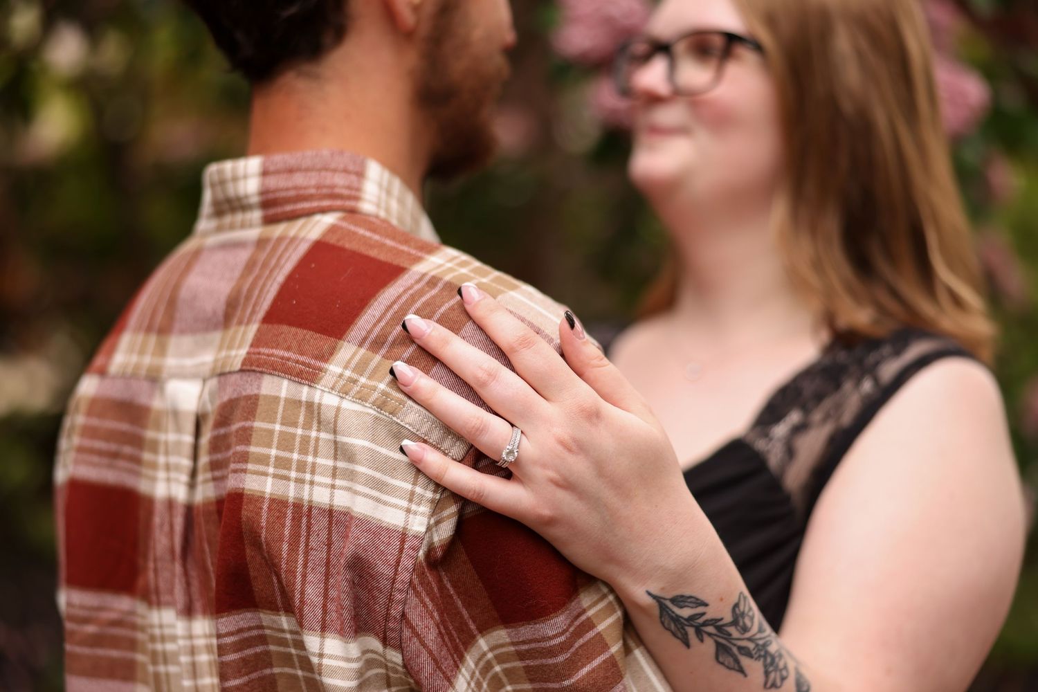 Engaged couple embracing outdoors, woman showing off her engagement ring with floral tattoo.