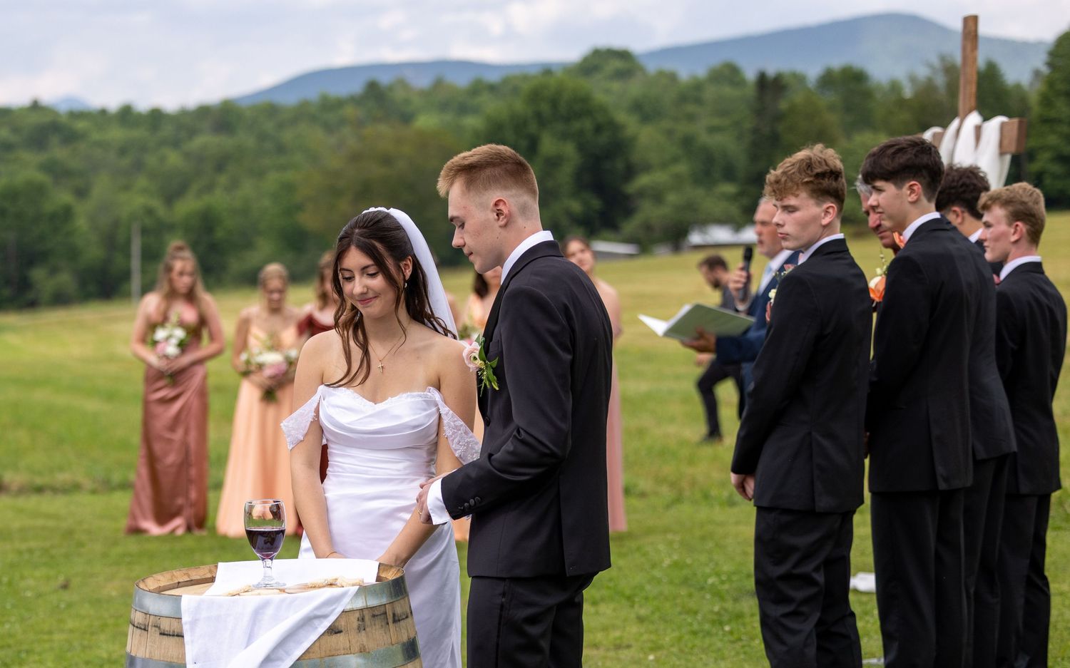 Bride and groom exchange vows at outdoor ceremony with green mountain backdrop and wedding party.