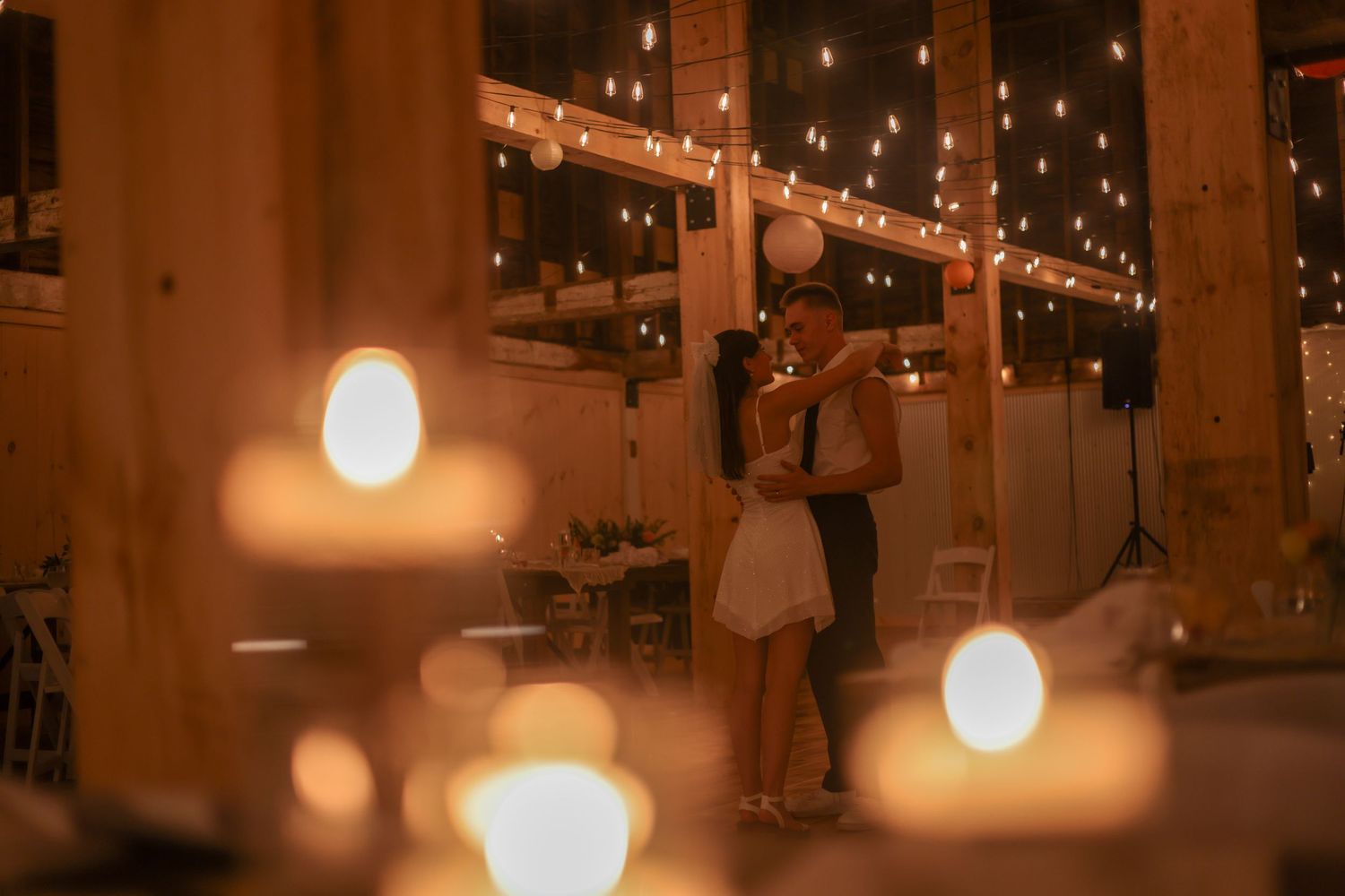 Romantic couple kissing during first dance surrounded by glowing candles at barn wedding reception.