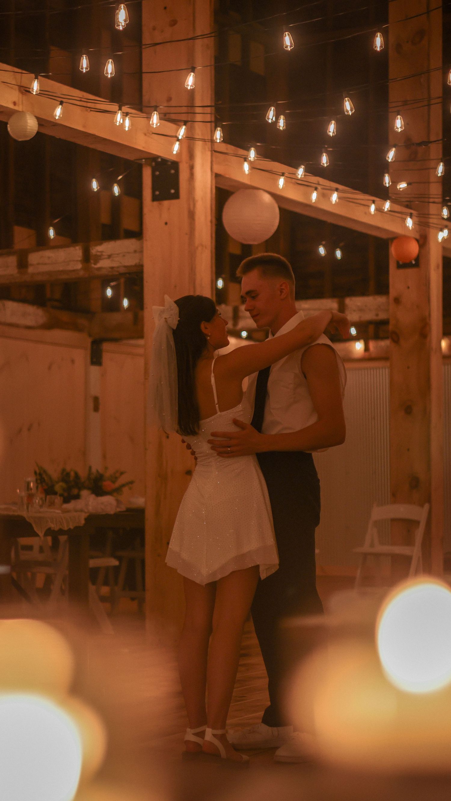 Bride and groom share first dance under warm string lights in a rustic barn wedding venue.