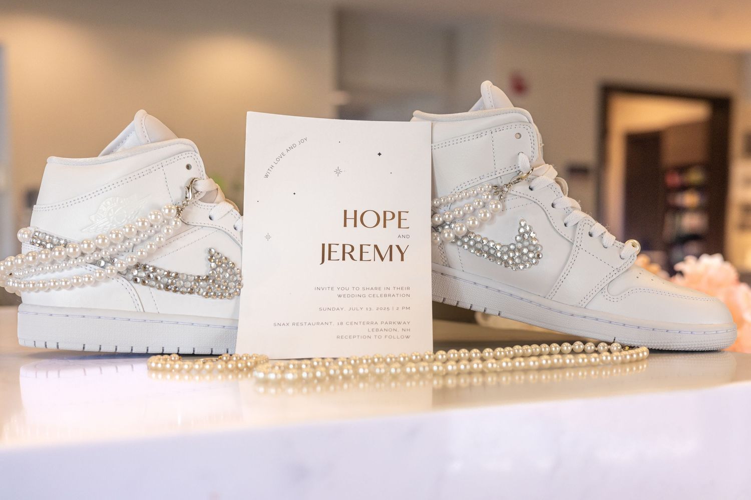 Custom pearl-embellished white Nike Air Jordan 1 sneakers beside Hope and Jeremy wedding invitation.