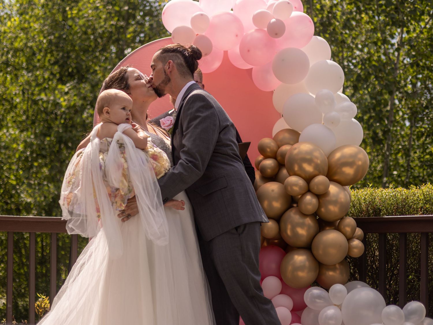 Couple sharing a kiss at outdoor wedding ceremony with pink and gold balloon arch, bride holding baby in white dress.