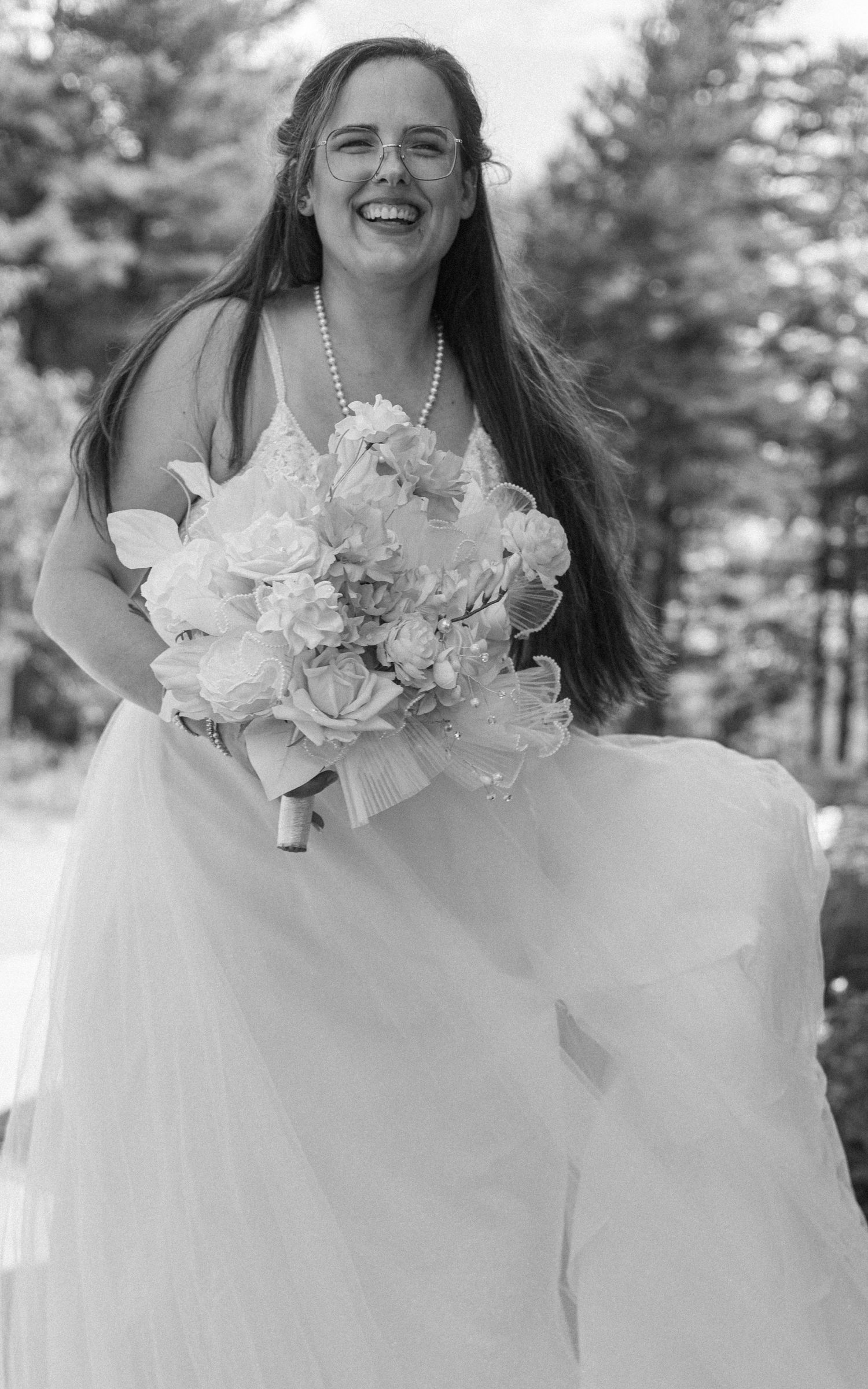 Joyful bride in white gown laughing while holding floral bouquet in black and white outdoor wedding photo.
