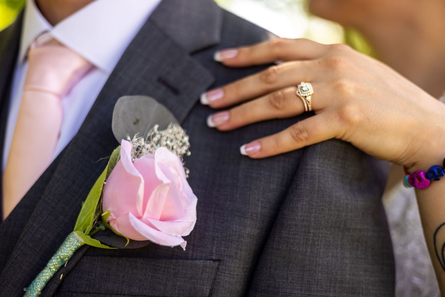 Close-up of groom's gray suit boutonniere with pink rose, bride's hand showing diamond wedding ring resting on chest.