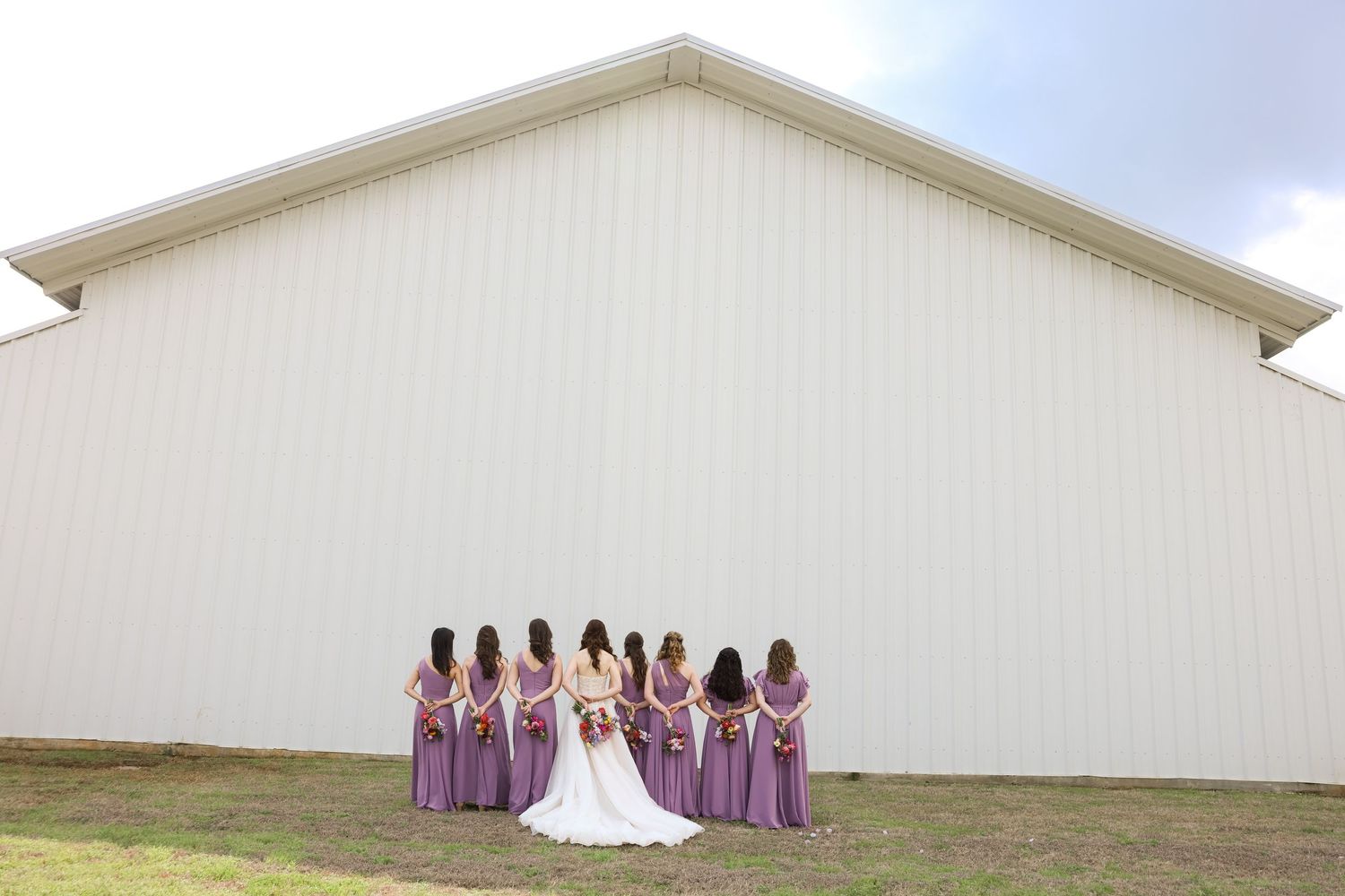 Bride and bridesmaids in lavender gowns standing facing a large white barn venue.