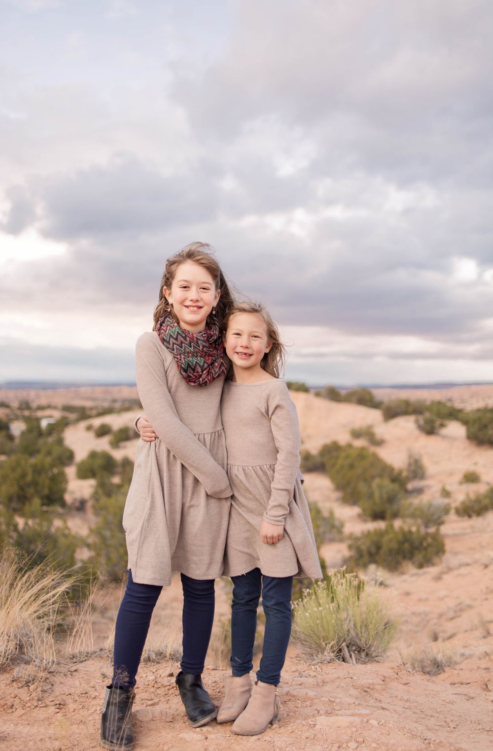 Two people in matching outfits embrace while standing in a desert landscape at sunset.