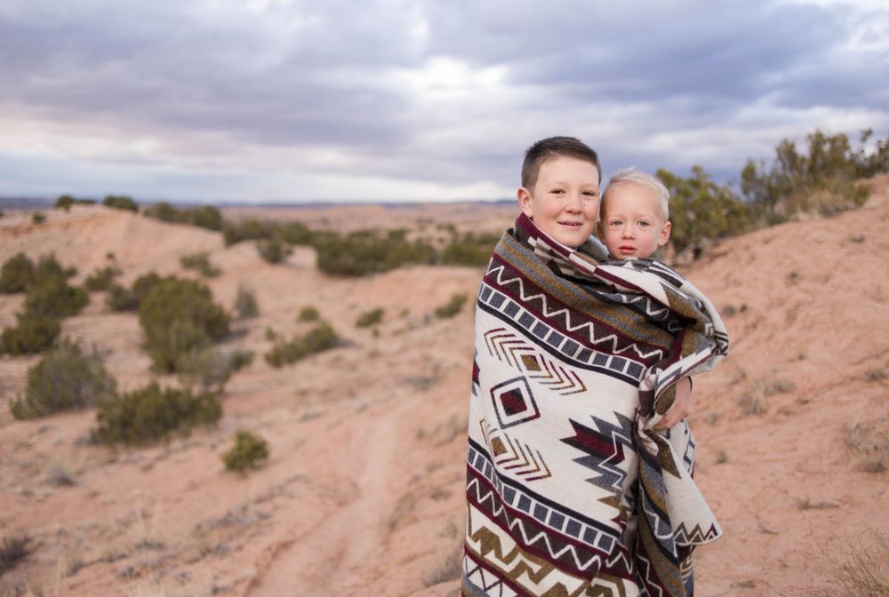 Two people wrapped in a patterned blanket stand together in a desert setting.